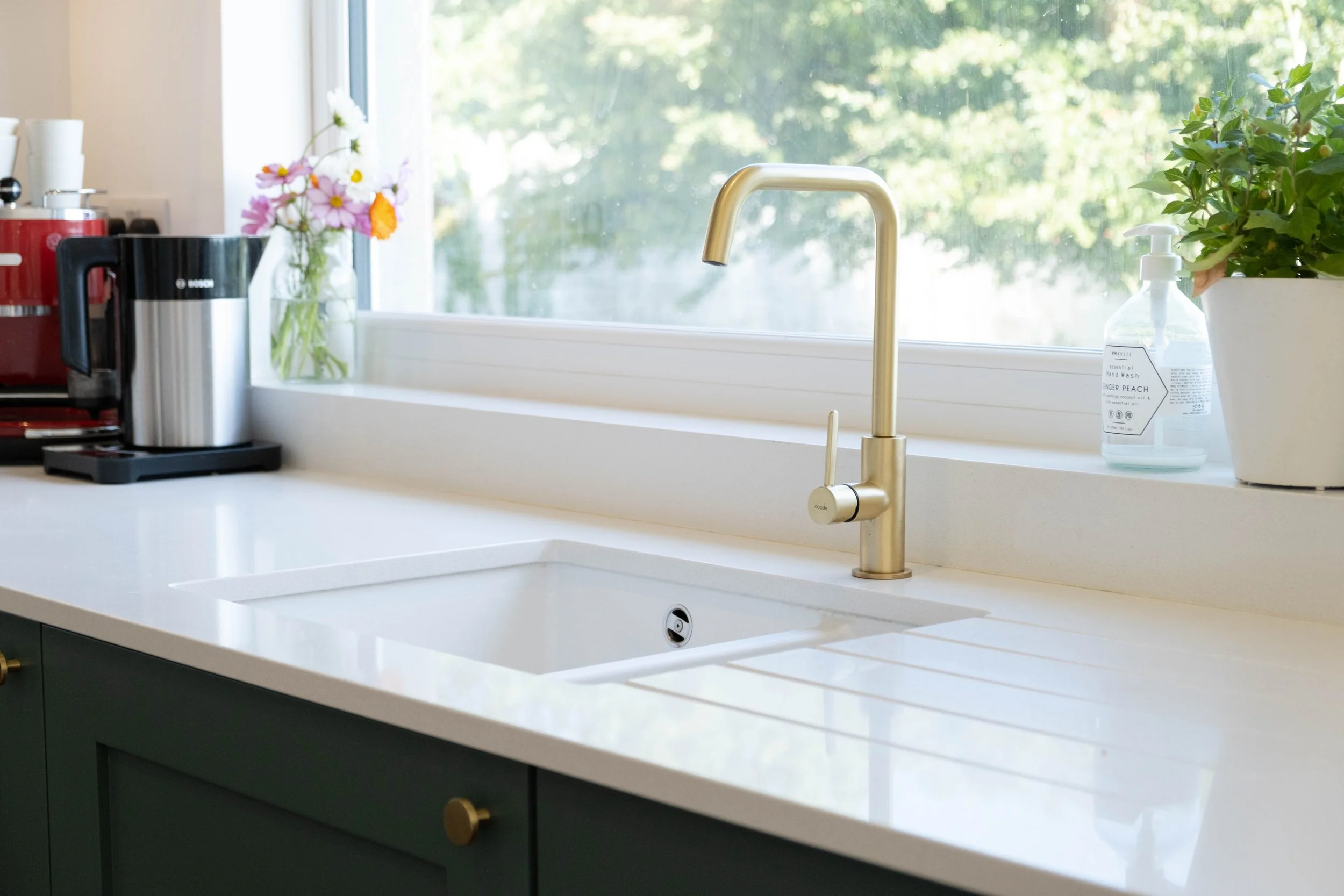 A modern kitchen sink with a gold faucet, a potted plant, a bottle of hand soap, a vase with pink and orange flowers, a coffee machine, and a red toaster, with a large window showing green trees outside.
