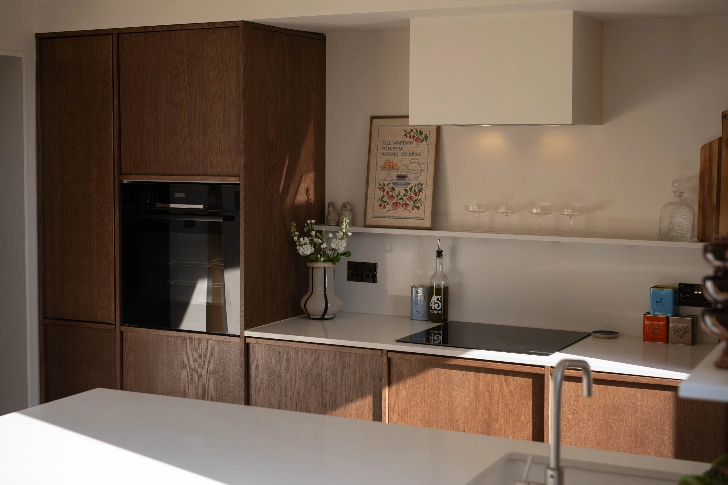 A modern kitchen with wooden cabinets, a white countertop, a framed cross-stitch picture, and decorative items including a vase of flowers and a glass water decanter.