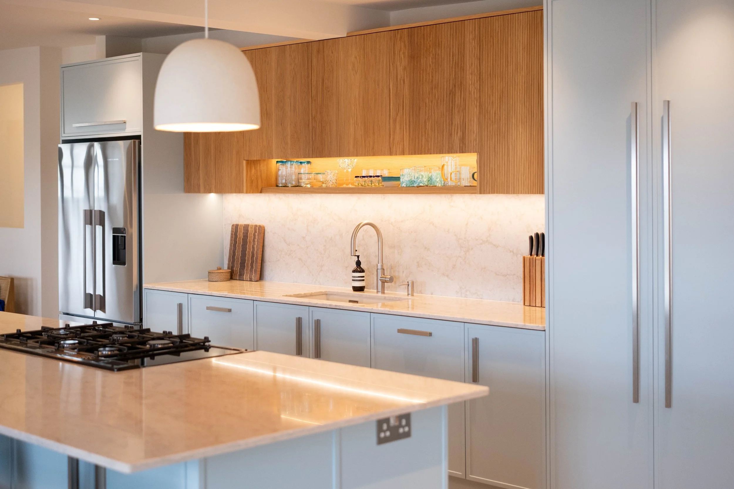 Modern kitchen with white countertops, light blue cabinetry, a stainless steel refrigerator, a built-in wooden shelf with glassware, and a hanging white pendant light.
