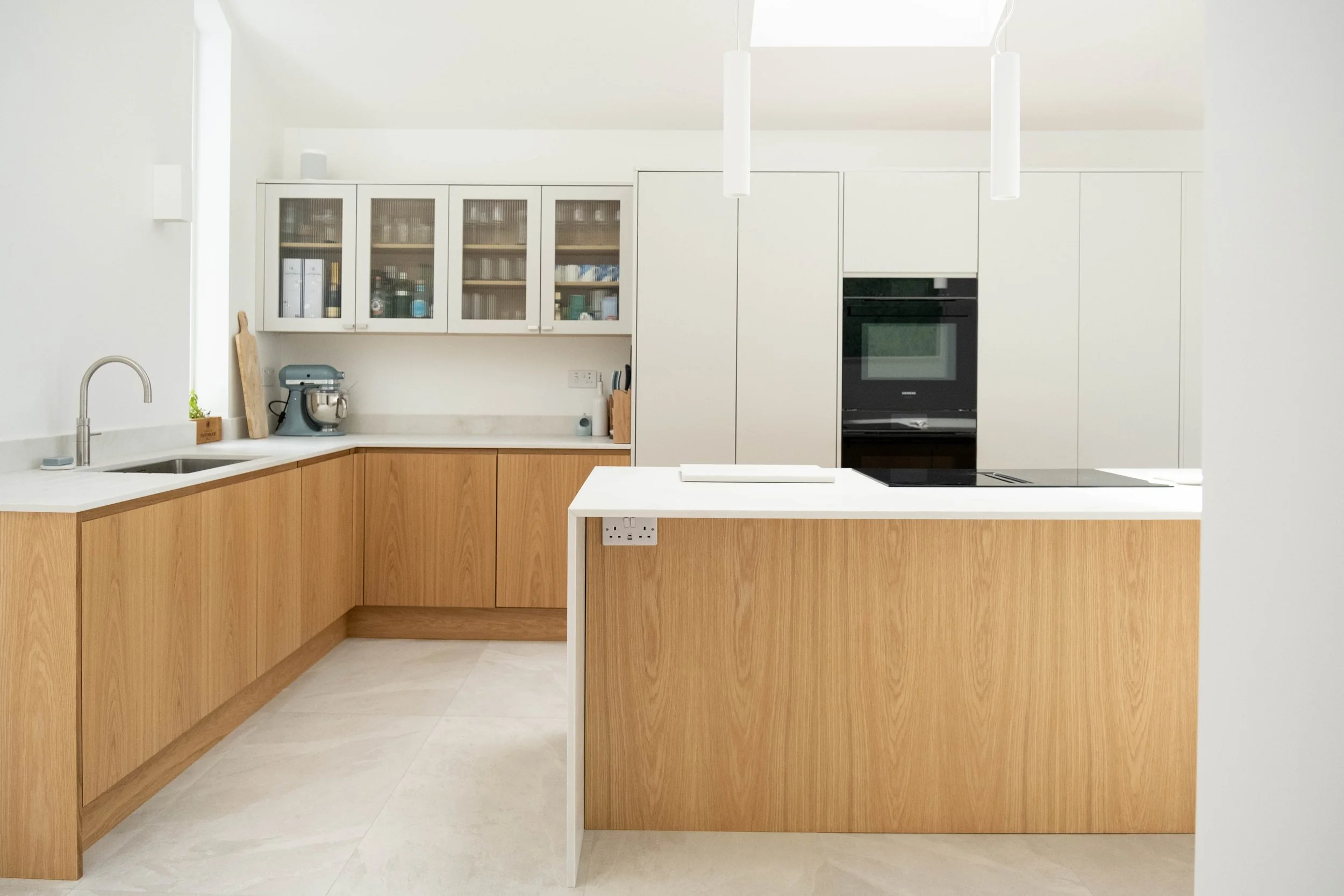 A modern kitchen with white upper cabinets with glass front, wooden lower cabinets, a white countertop, a sink, a mixer, and a black oven. Two white pendant lights hang from the ceiling.
