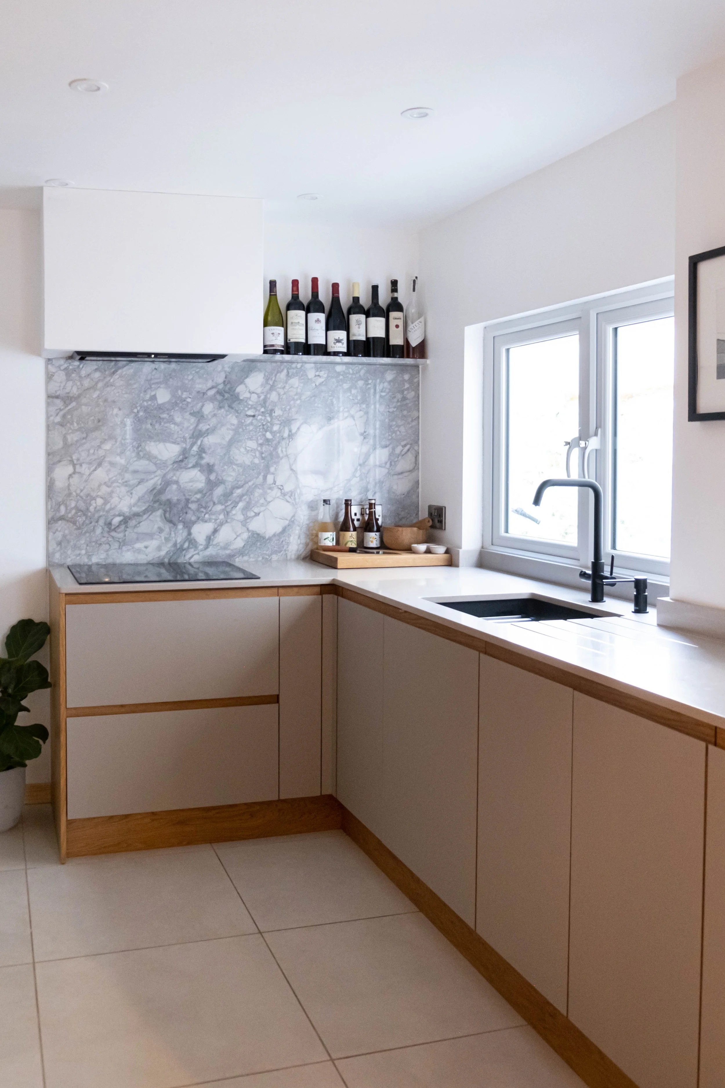 A modern kitchen with a white marble backsplash, beige cabinetry with wooden trim, a black sink faucet, a window with a view outside, and bottles of wine on the open shelf above the backsplash.