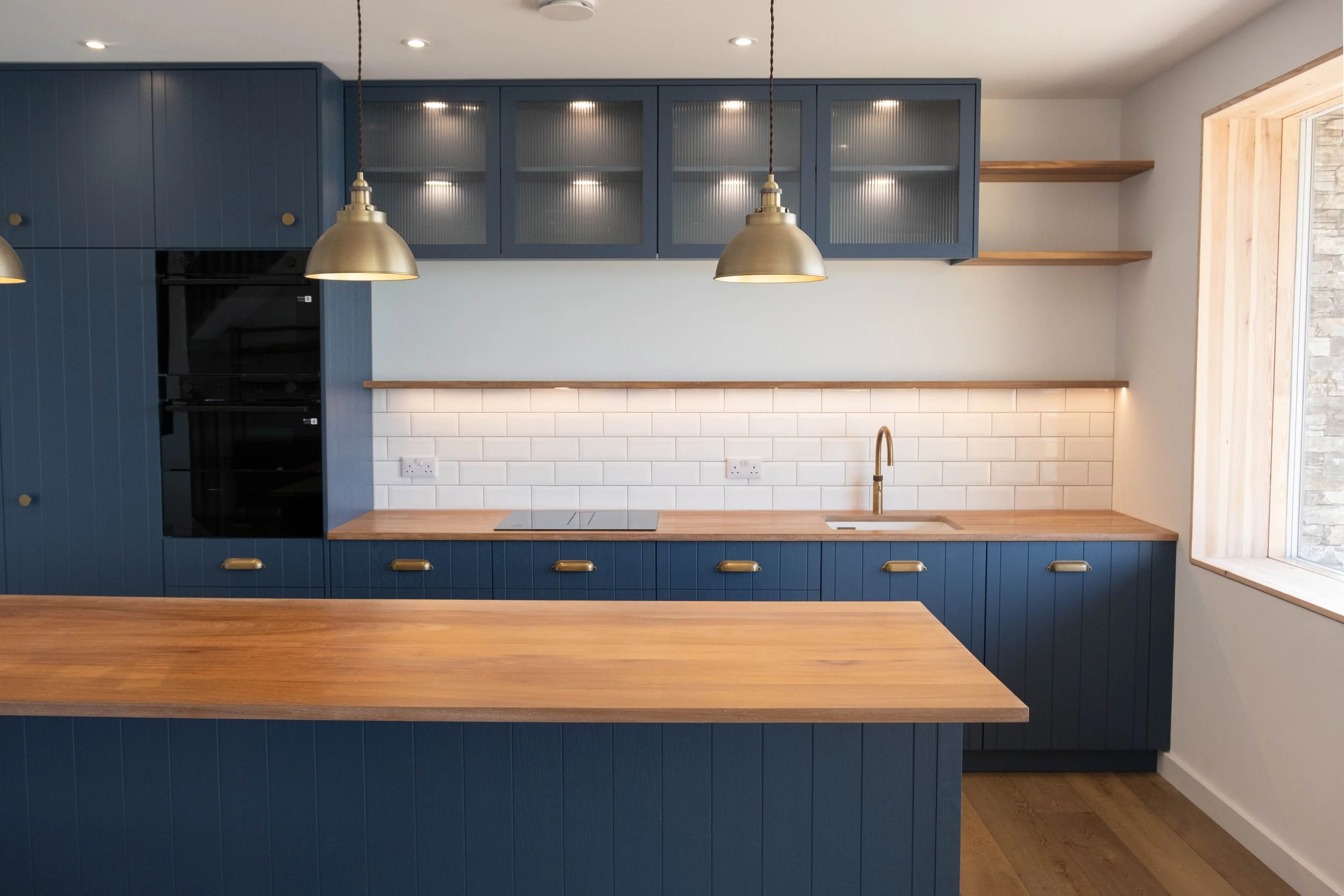 Modern kitchen with blue cabinets, open shelves, a white tiled backsplash, a wooden countertop, and two brass pendant lights hanging above the counter.