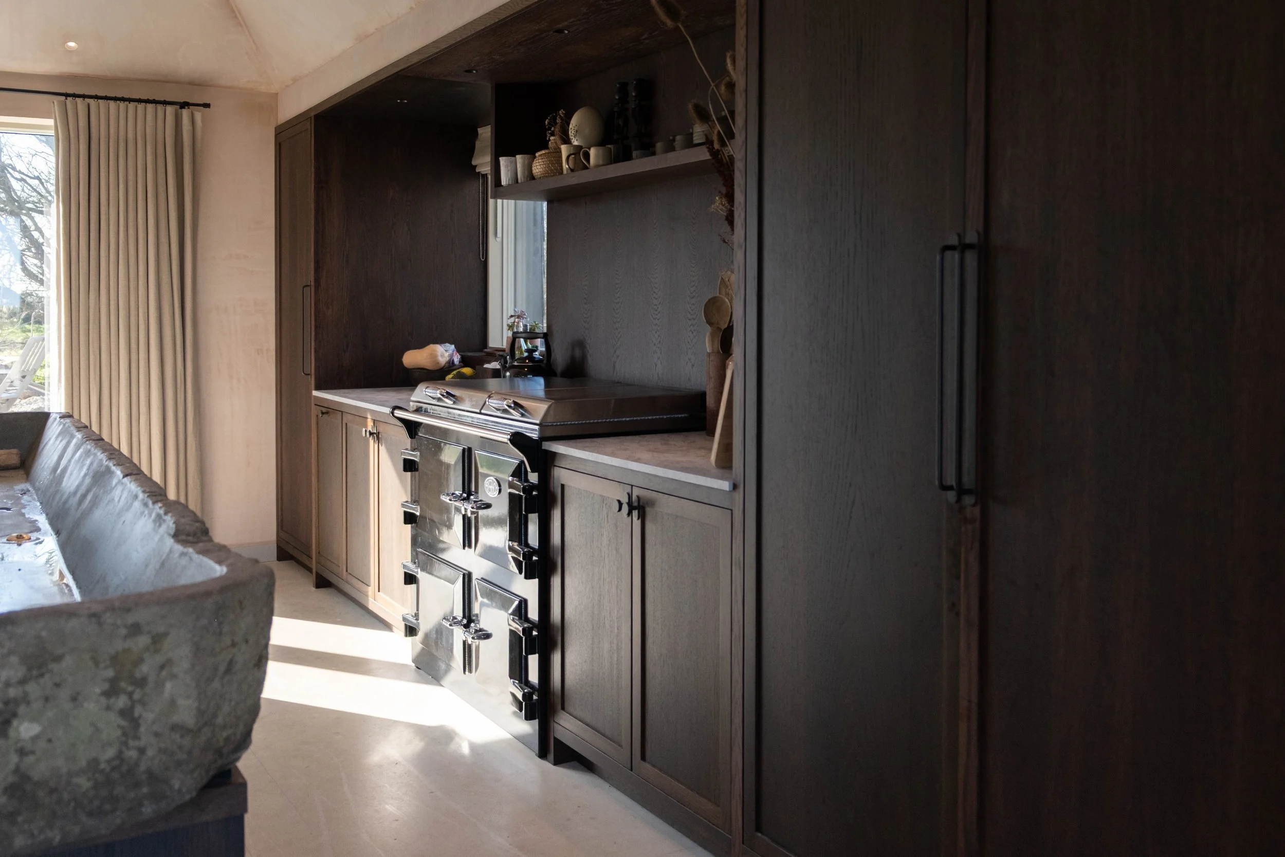 Interior view of a kitchen with dark wood cabinets, a stainless steel stove, and light-colored countertops. There is a small window above the counter, and a section of dining table with a stone or concrete edge is visible on the left side. A large sl