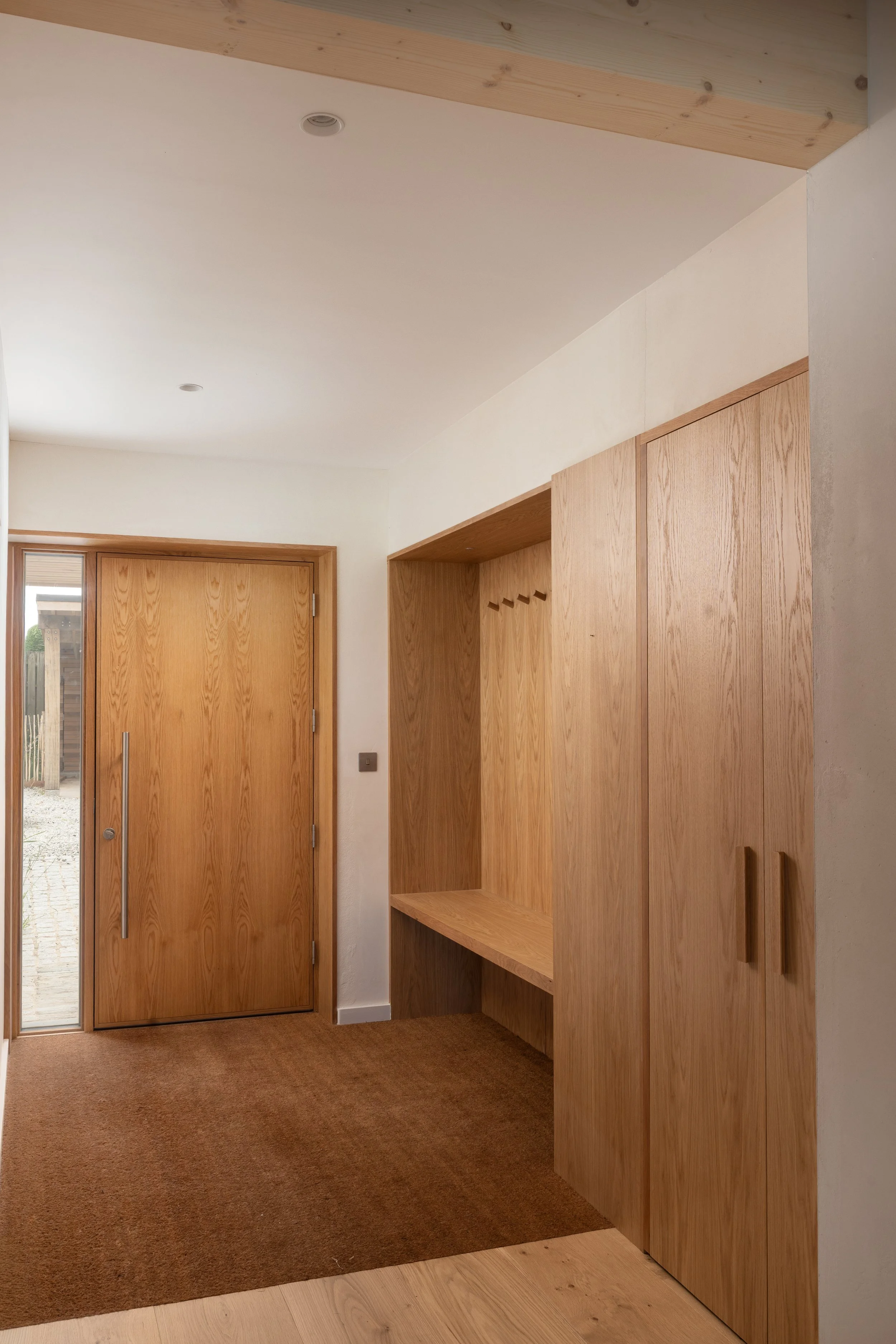Interior view of a wooden entryway with a closed wooden door on the left, a small section of glass window, a built-in wooden storage unit with hooks, and a brown carpet on the floor.