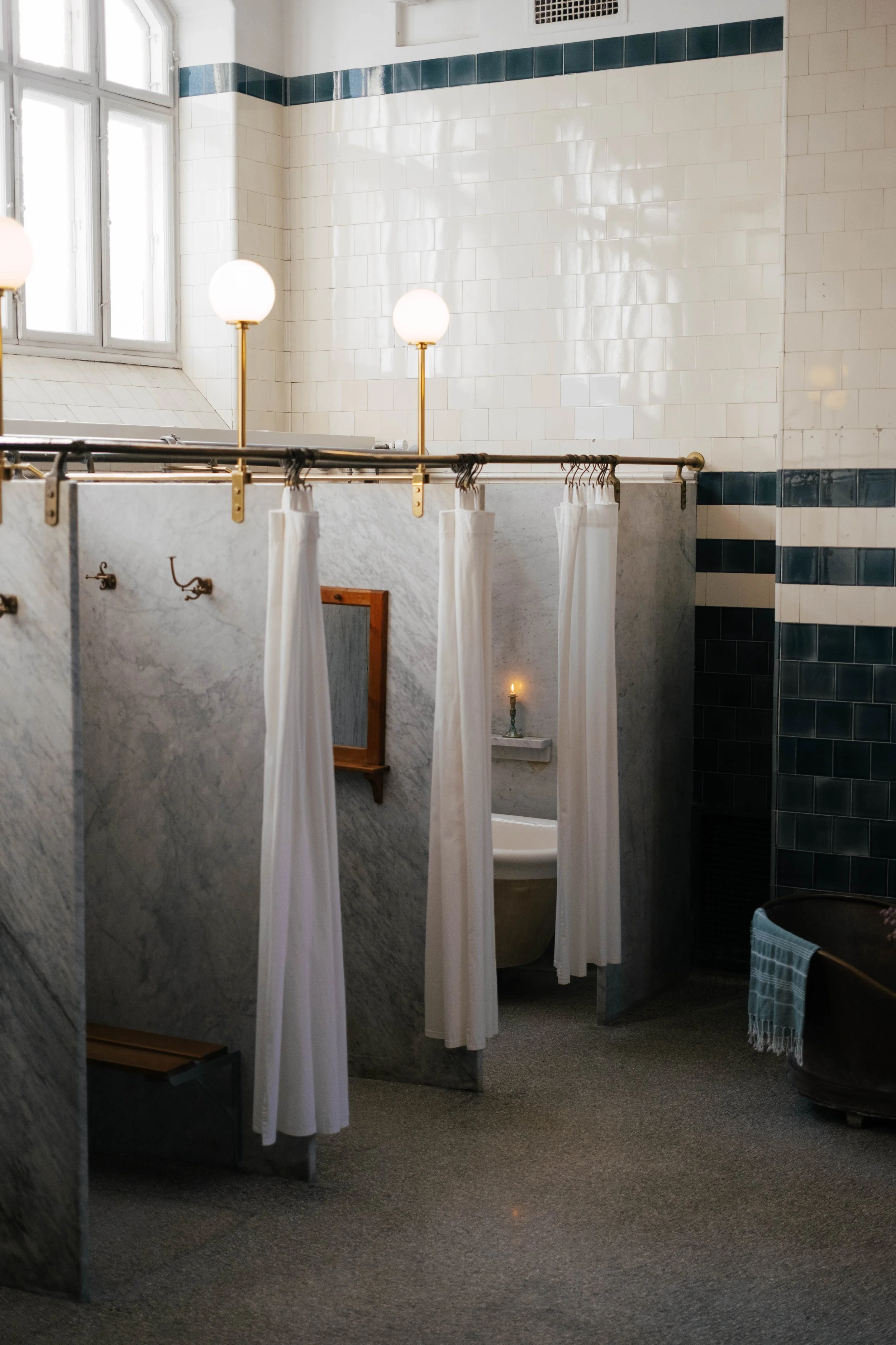 A changing room with white tiled walls accented with blue tiles near the top. There are three light fixtures with round bulbs on brass stands, and a window with multiple panes letting in natural light. A vanity mirror with a wooden frame and a small 