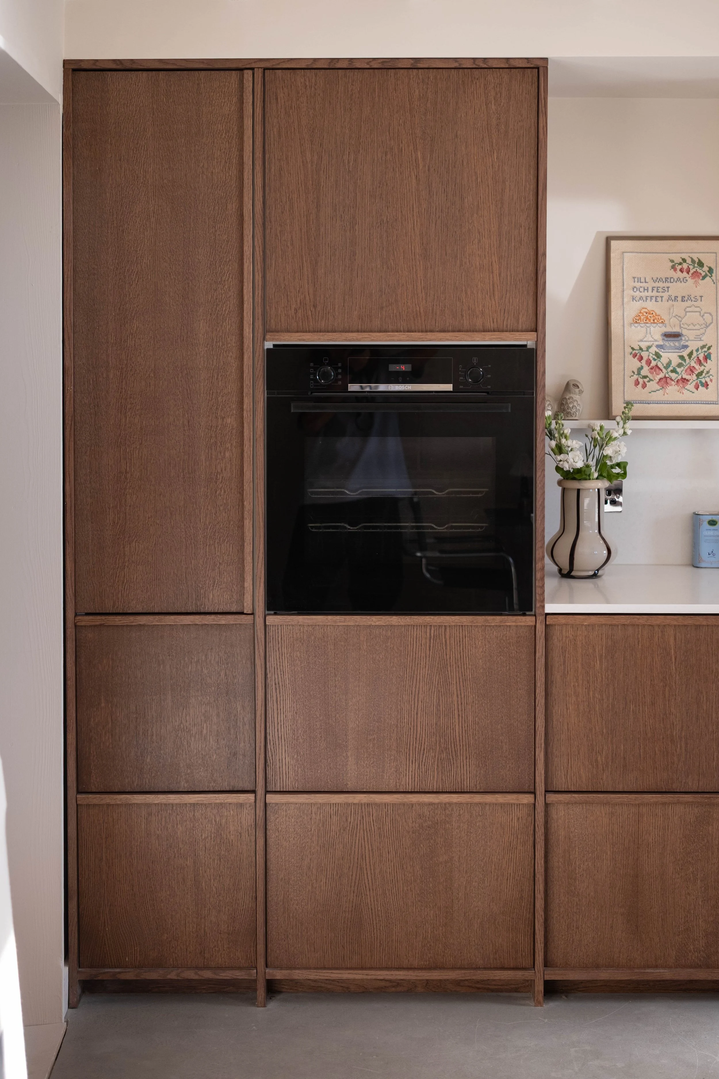 A built-in wooden cabinet with a black oven in the center, a white wall with framed artwork, a vase with white flowers, and a small appliance on the right side.
