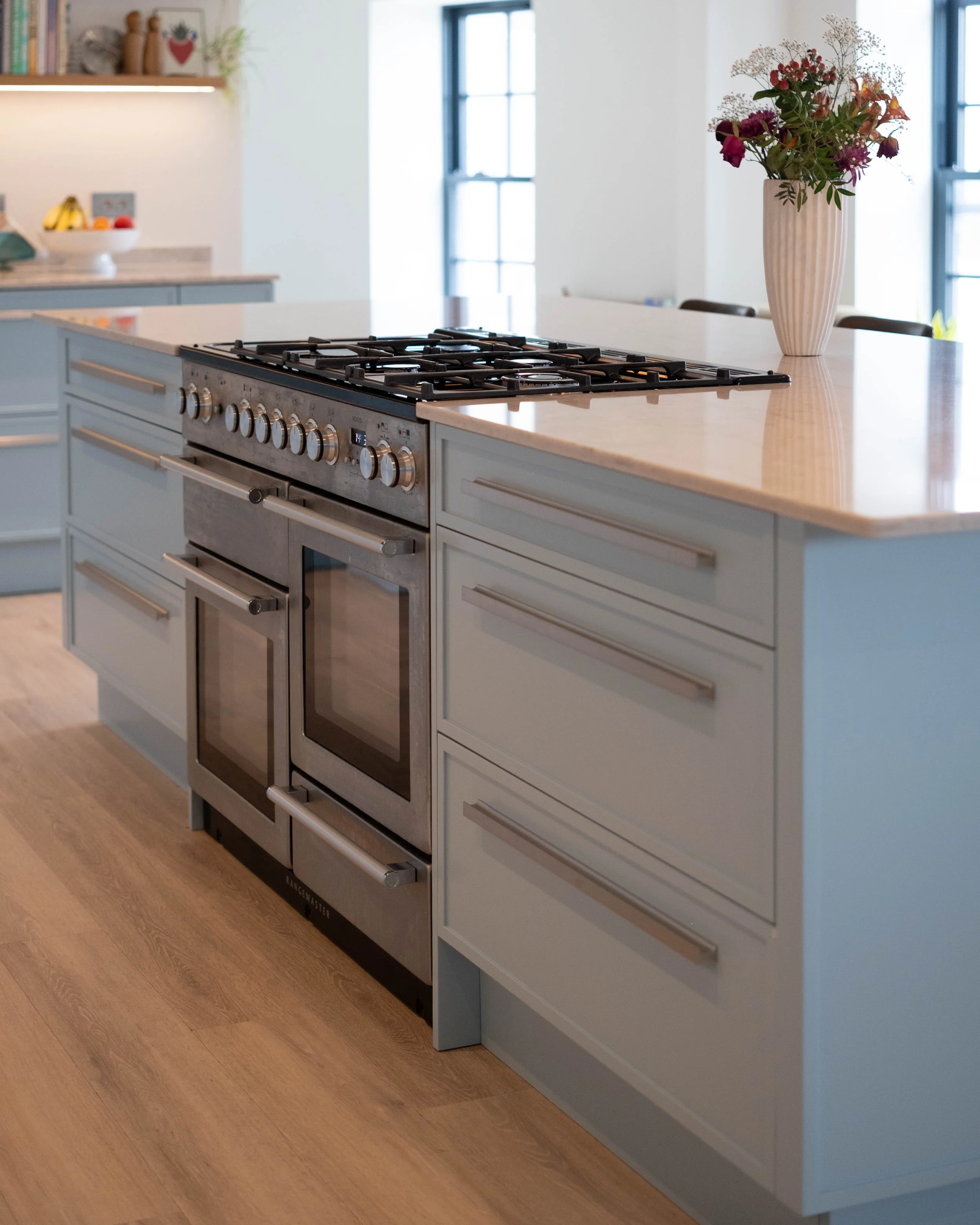 A modern kitchen island with a built-in stainless steel oven and stovetop, light blue cabinets, a marble countertop, a vase with flowers, and a window in the background.