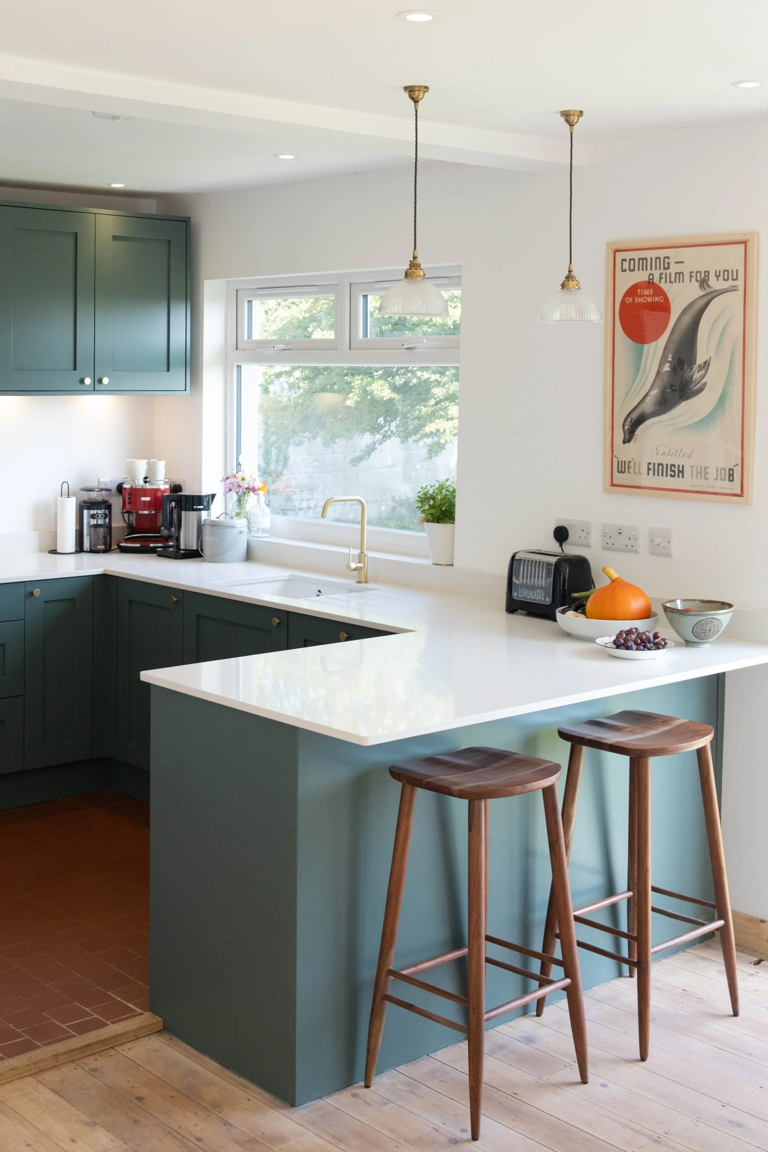 A kitchen with green lower cabinets and a white countertop, with two wooden bar stools, a pumpkin, a bowl of grapes, a toaster, and coffee-making appliances on the counter. There is a window with a green outdoor view and a decorative wall poster.
