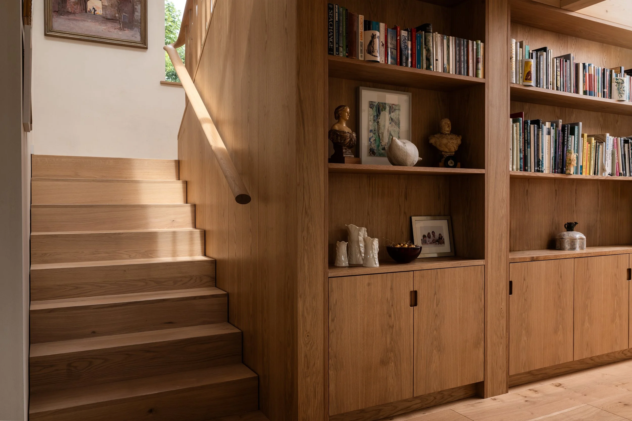 A wooden staircase next to a built-in bookshelf with various books, decorative items, and framed photos inside a home.