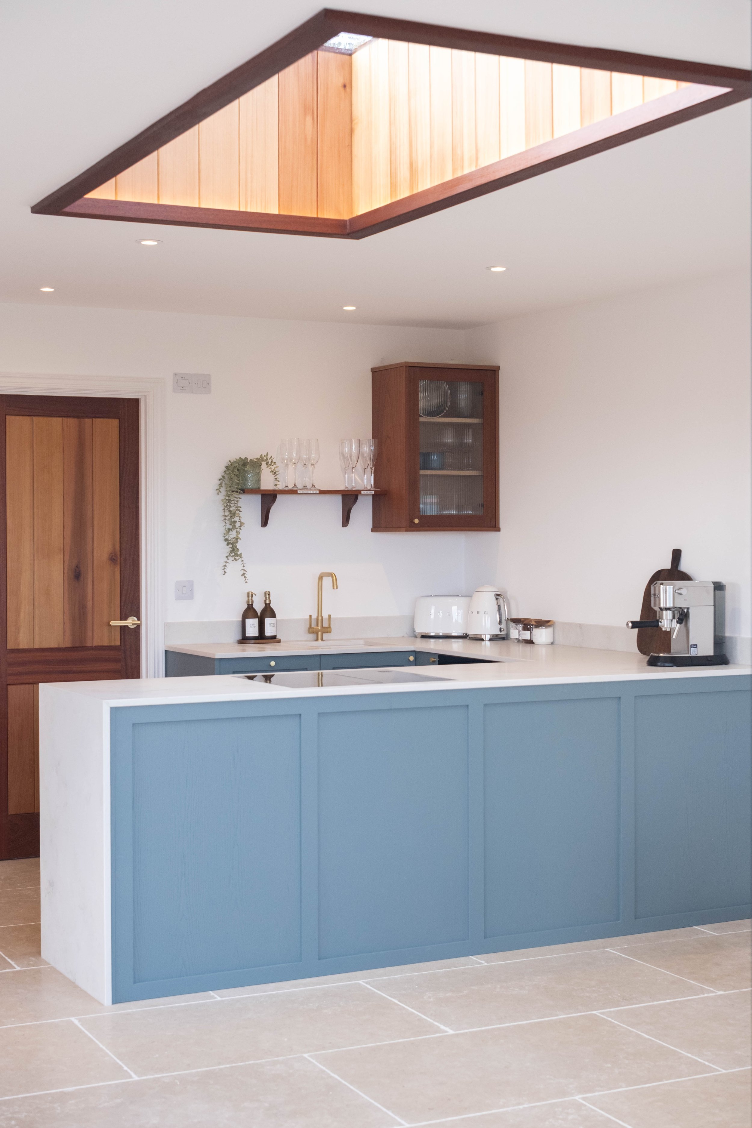 A modern kitchen with a blue kitchen island, white countertop, wooden door, and a ceiling skylight with wood paneling. The kitchen features small appliances, glasses, and decorative bottles.