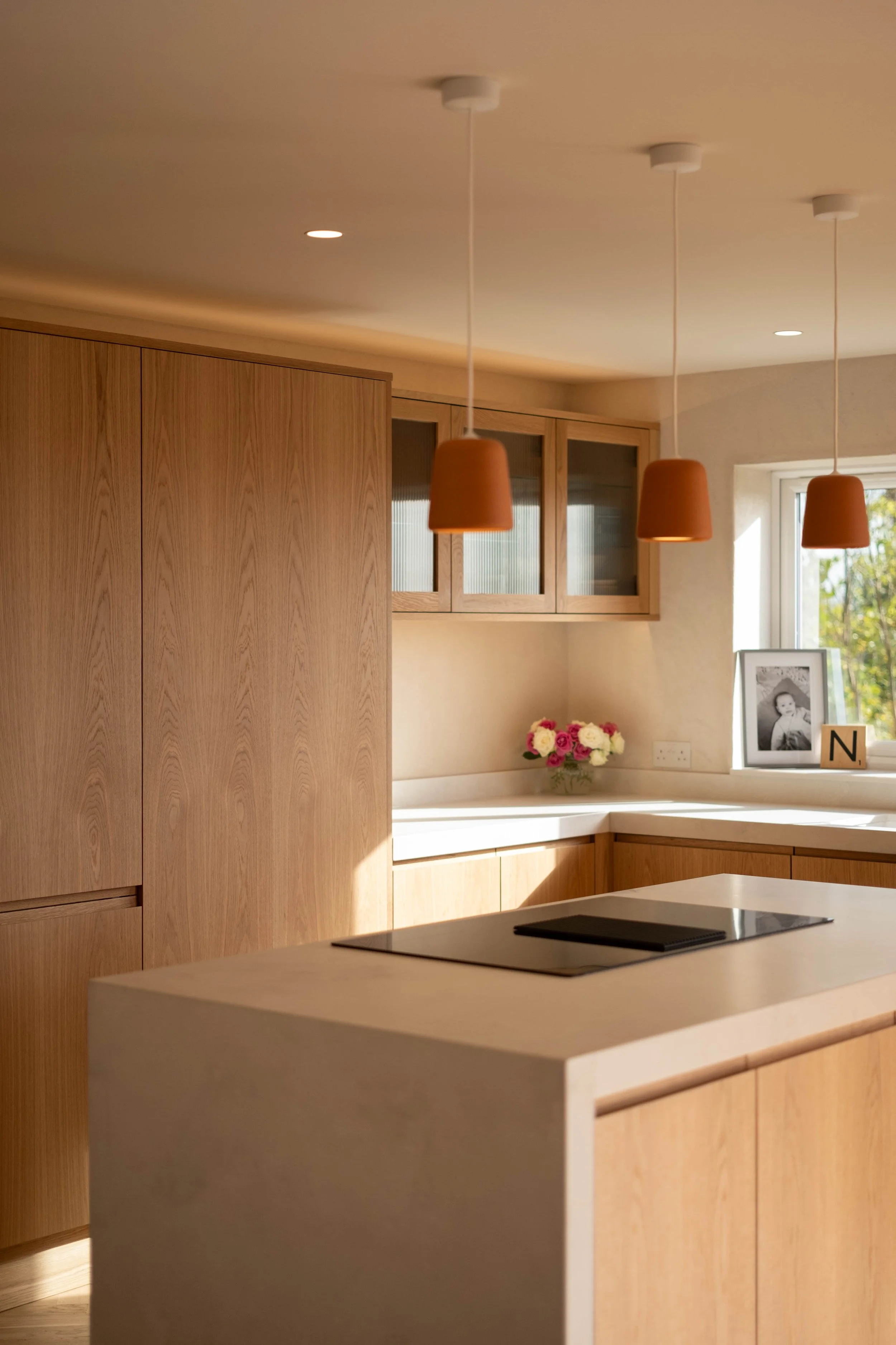 Modern kitchen with wooden cabinets, a white countertop, three hanging terracotta pendant lights, a window with natural light, a framed photo, a bouquet of flowers, and a black cooktop on the island.