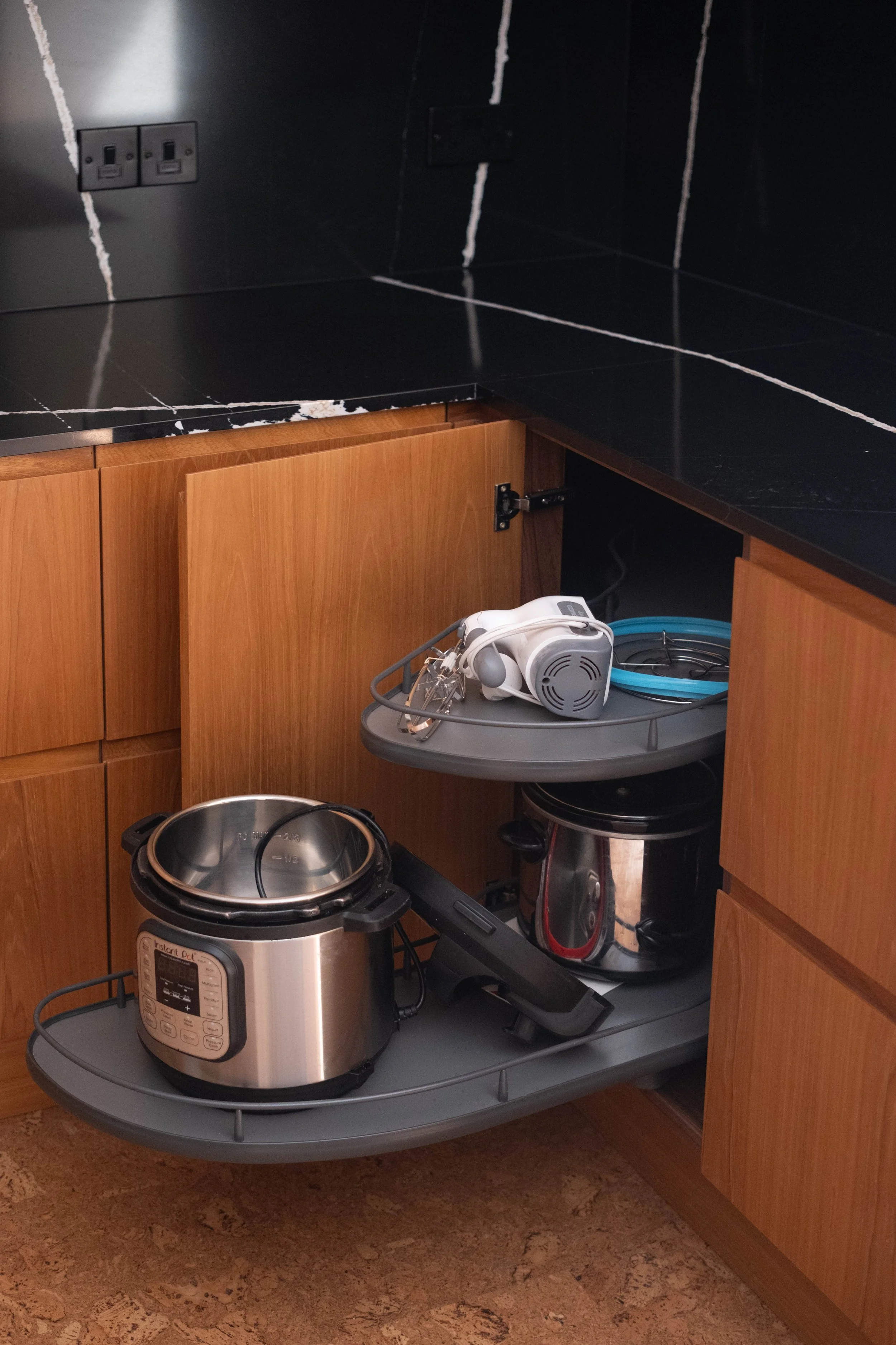 The kitchen has black marble countertops and wooden cabinets. A white kitchen appliance, possibly a food processor or stand mixer, is placed on a gray countertop extension. There is a slow cooker or pressure cooker on the floor and a vacuum cleaner p
