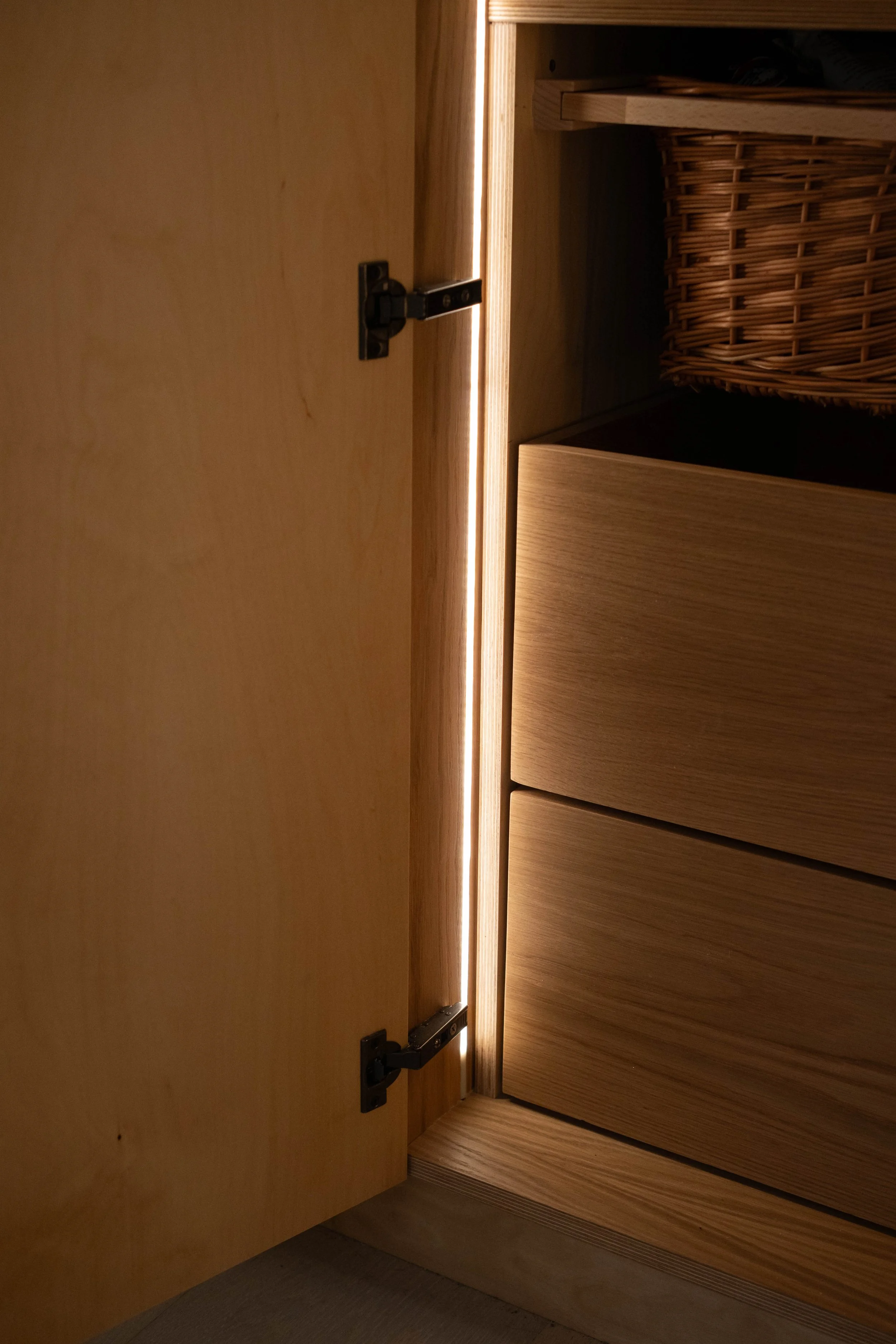 Open wooden cabinet door revealing shelves inside, including a wicker basket, with warm interior lighting.