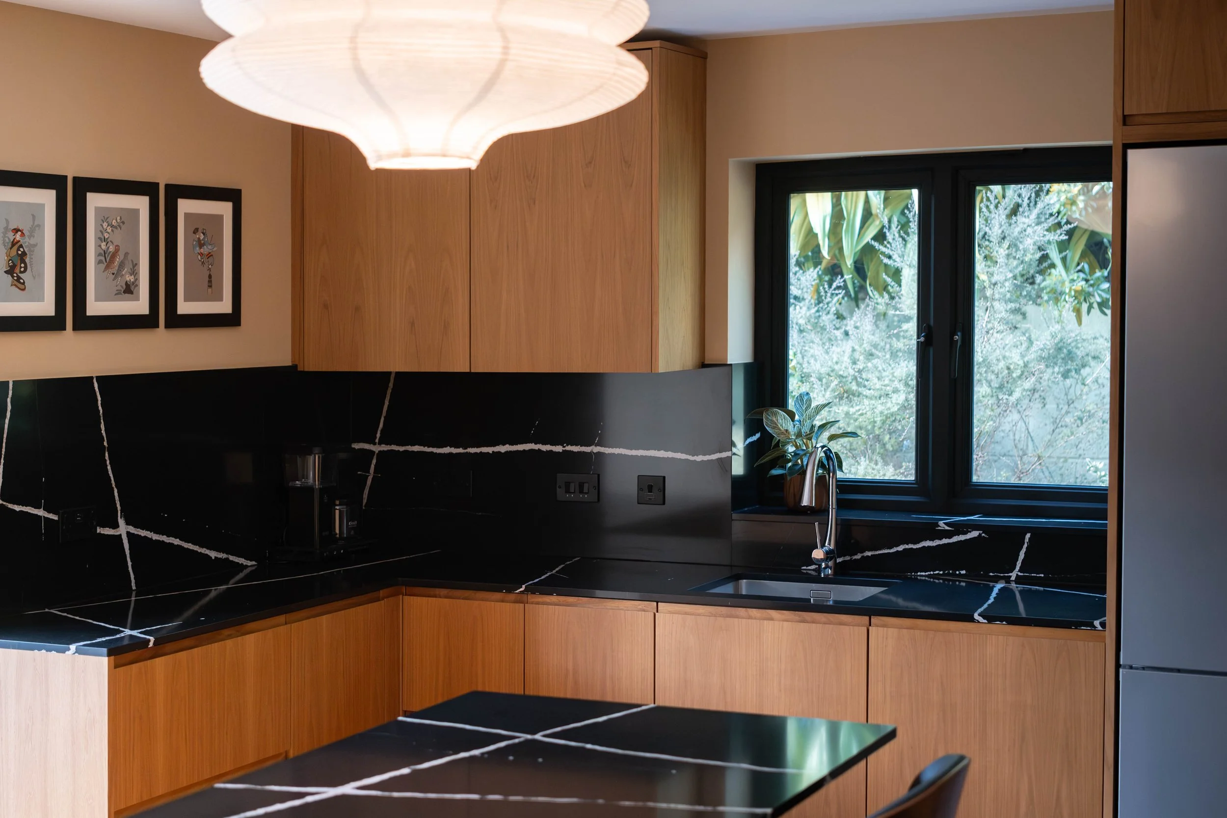 Modern kitchen with black marble countertops featuring white veining, wooden cabinets, a black window overlooking greenery, and framed artwork on the wall. A white paper lantern-style ceiling light hangs above.