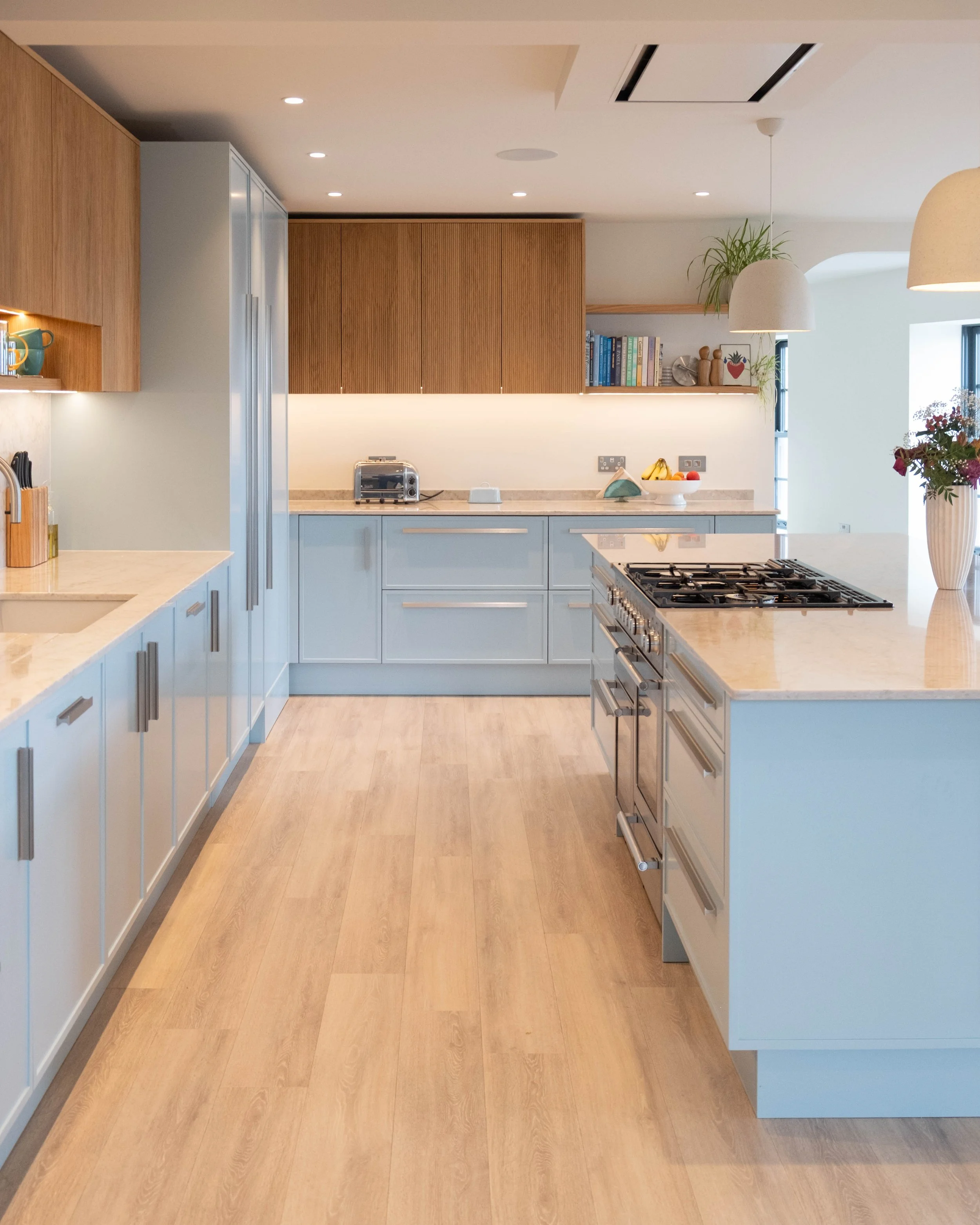 Modern kitchen with light blue cabinets, wooden upper cabinets, a marble countertop, built-in appliances, a bowl of fruit, and decorative items including books, plants, and vases.