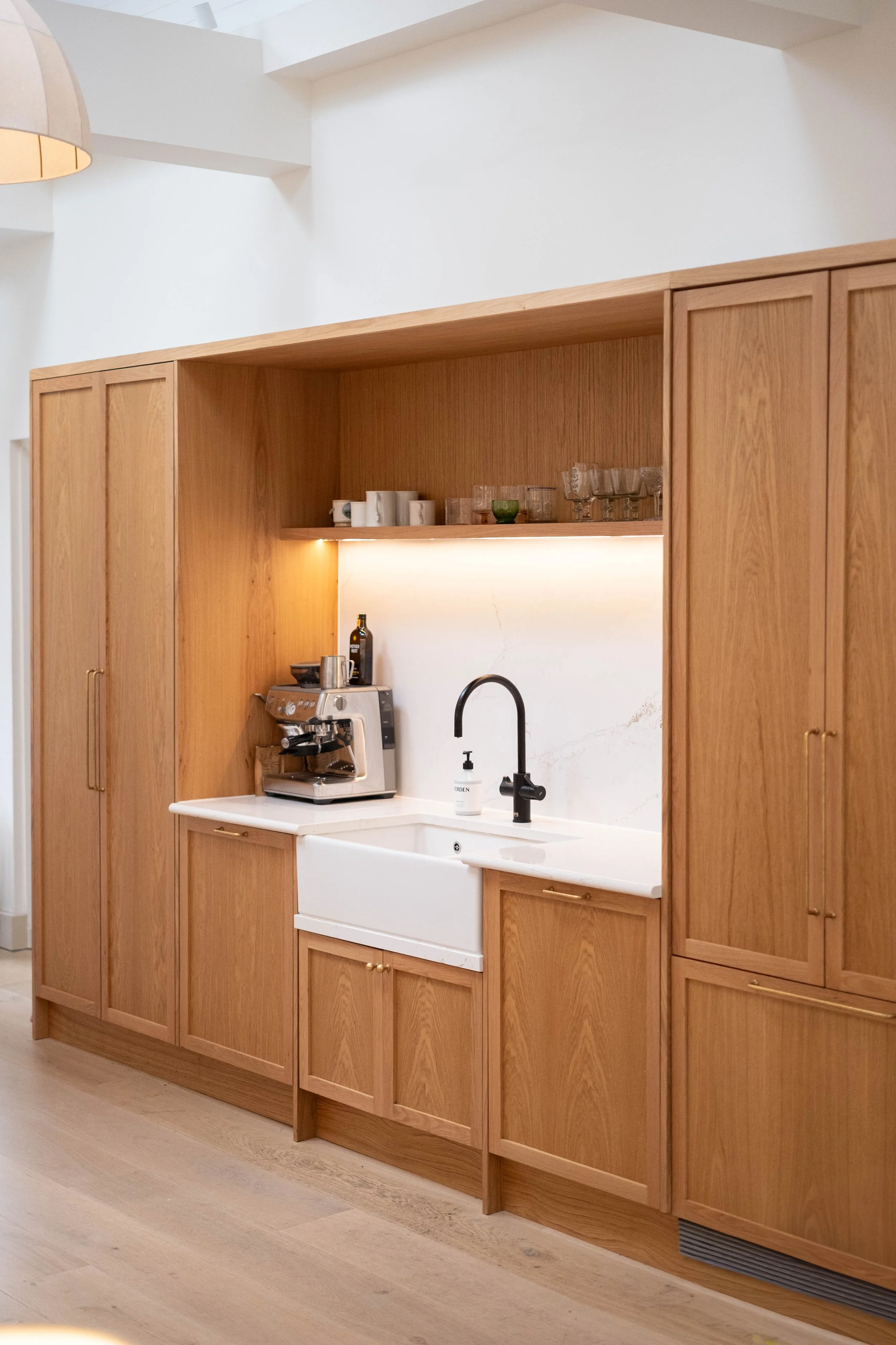 Kitchen area with wooden cabinets, a coffee machine, a black faucet, and a white farmhouse sink. Open shelving with mugs, glasses, and a small green bowl. Light-colored wood flooring and a white wall with marble accents.