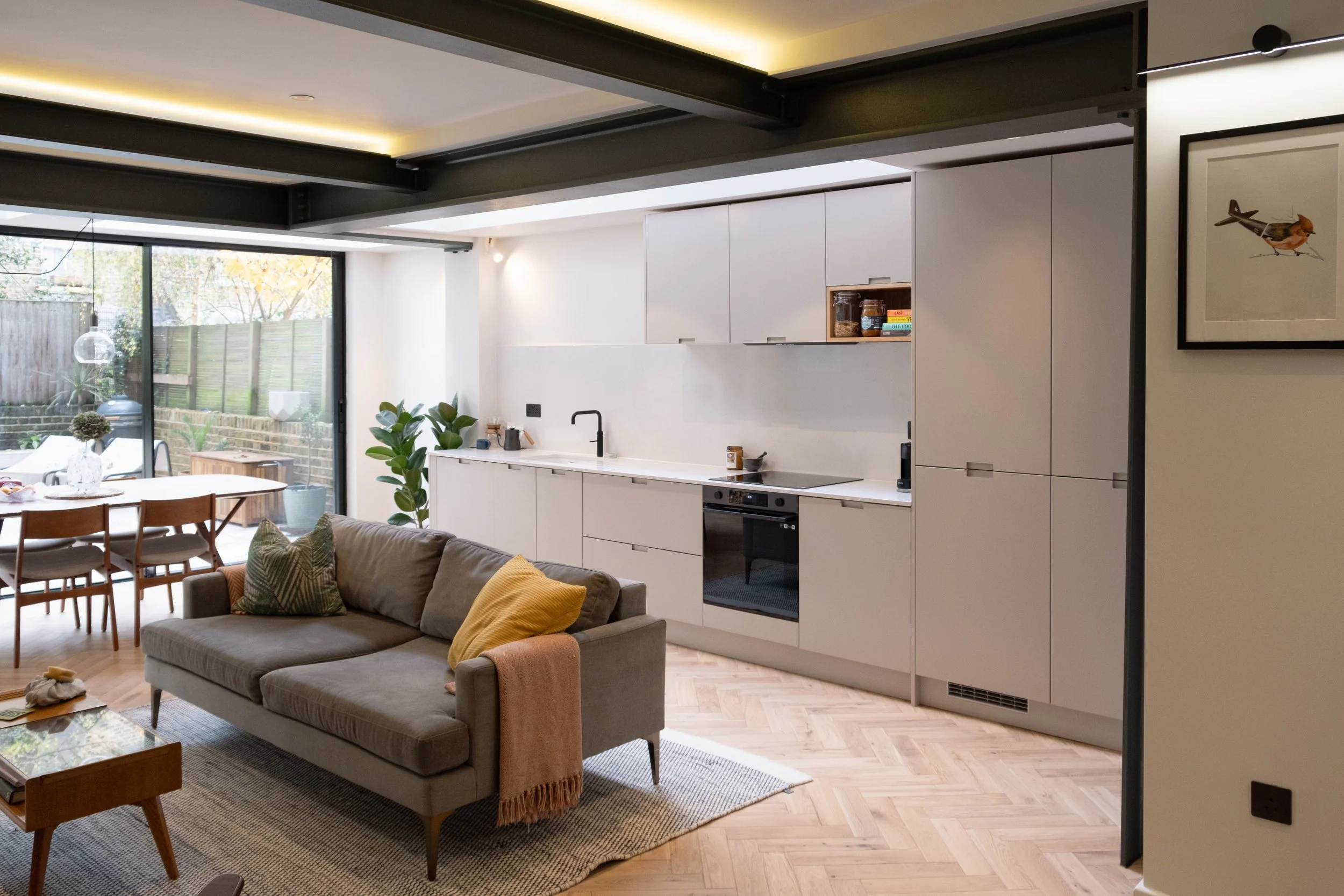 Modern open-plan living room and kitchen with large sliding glass doors, wooden flooring, a sofa with cushions, a dining table with chairs, kitchen cabinets, and plants.