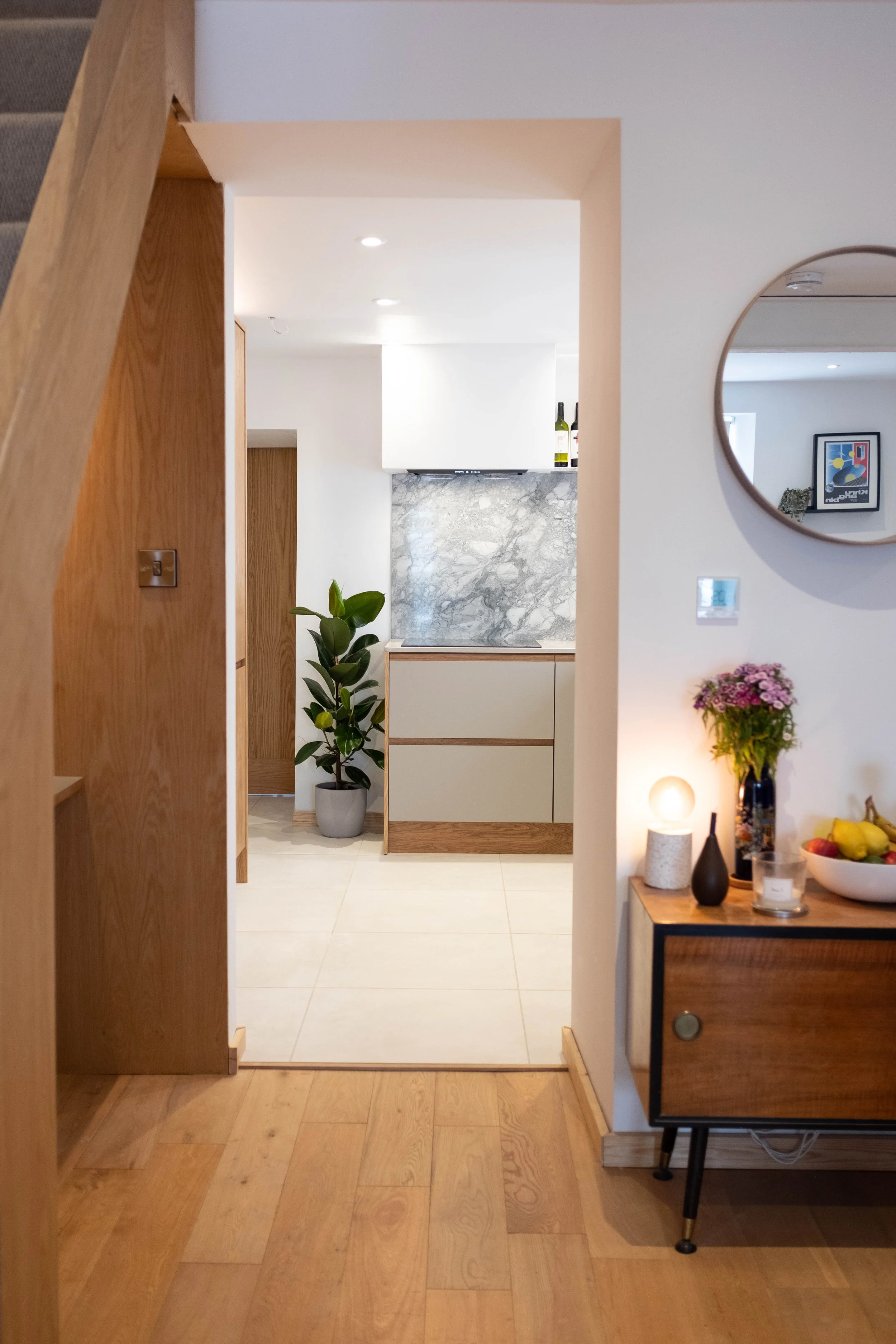A modern living space with a view into the kitchen area. The foreground includes a wooden partition, with a partial view of a TV on a black stand with gold-tipped legs, placed on a wooden floor. To the right, there is a small table with a vase of pur
