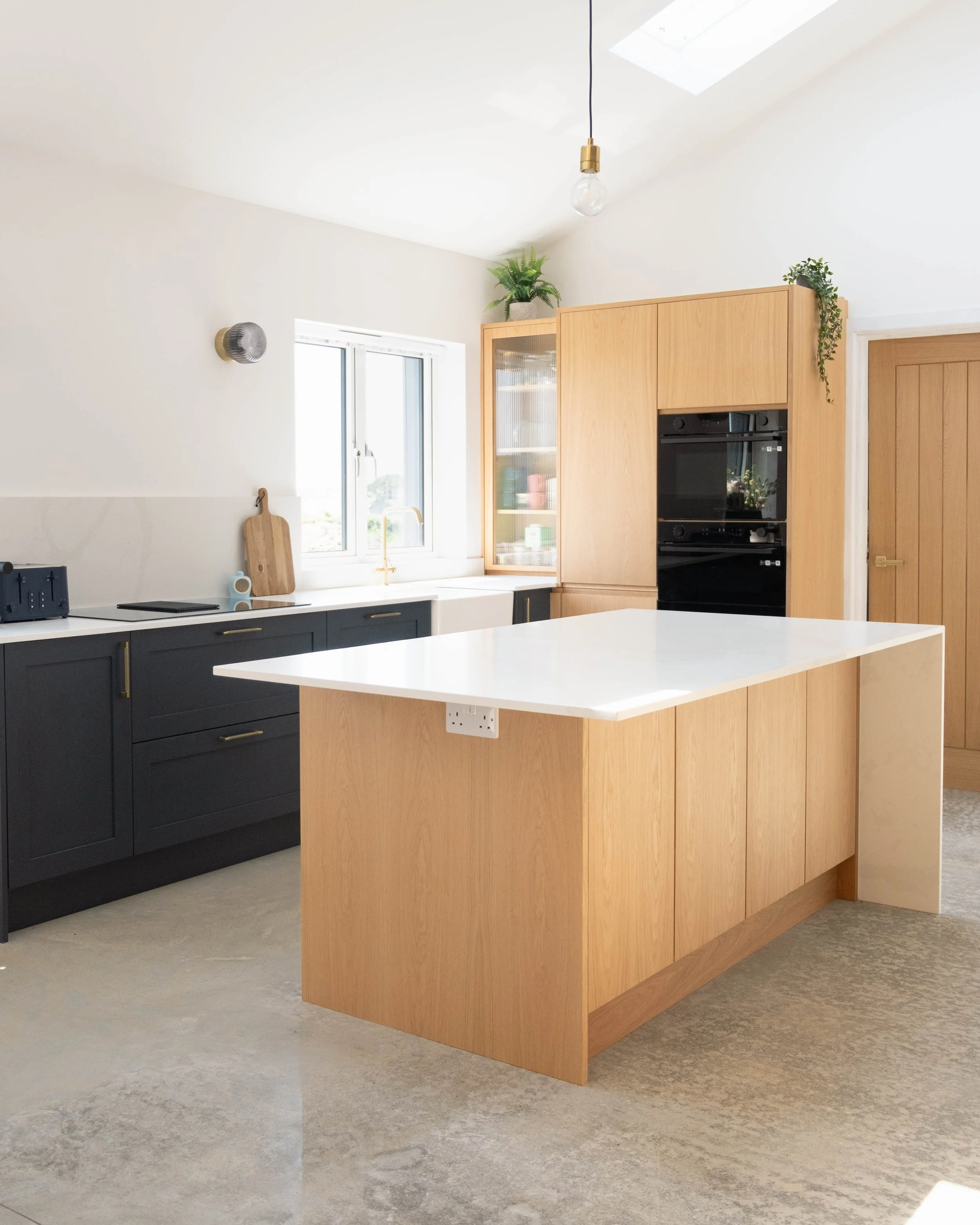 Modern kitchen with a large wooden island, black cabinets, a white countertop, a window with a view, and a hanging light bulb fixture.