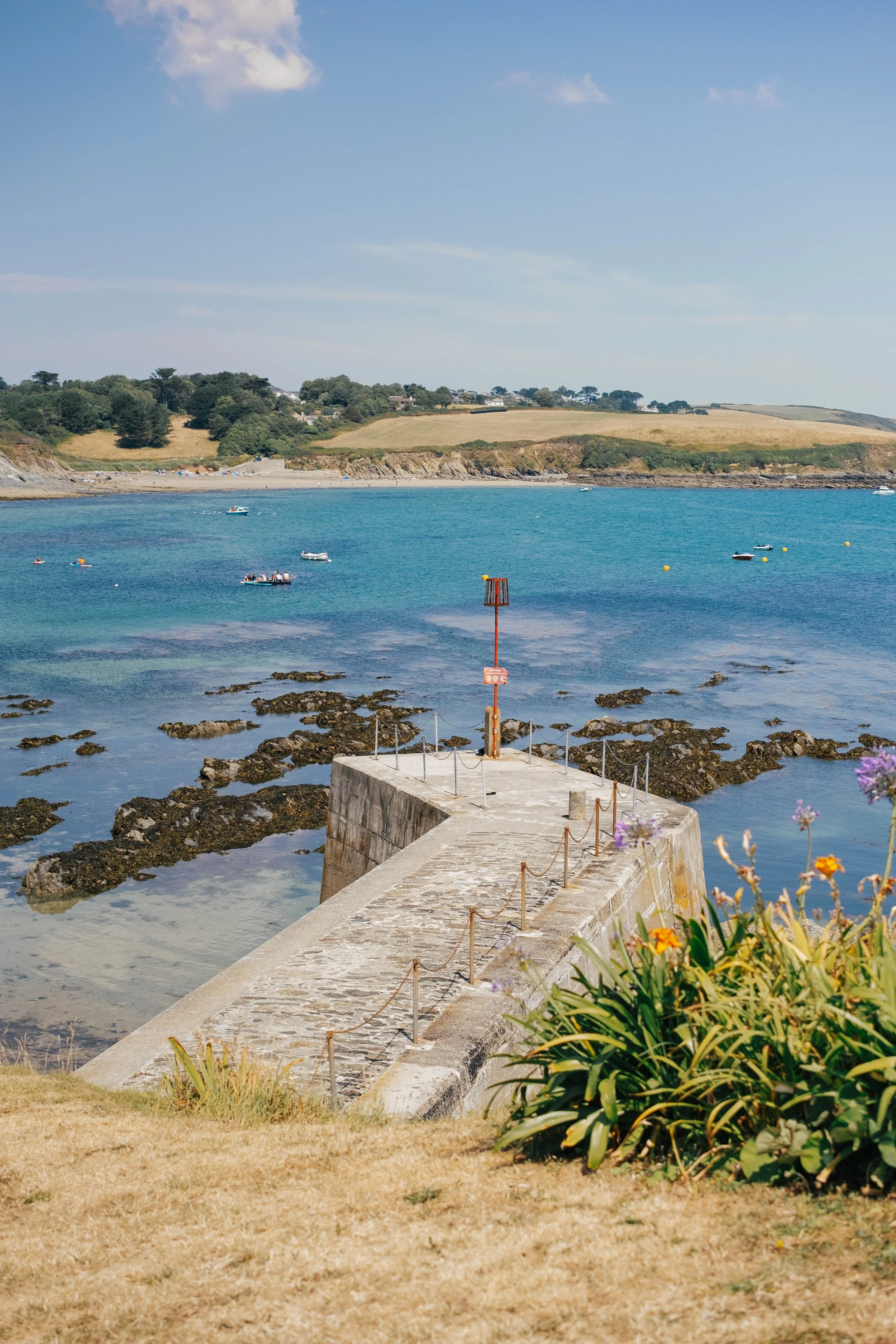 A coastal scene with a stone pier extending into calm blue water. Small boats are floating on the water, while a grassy hillside with trees and houses is in the background. The sky is mostly clear with a few scattered clouds.