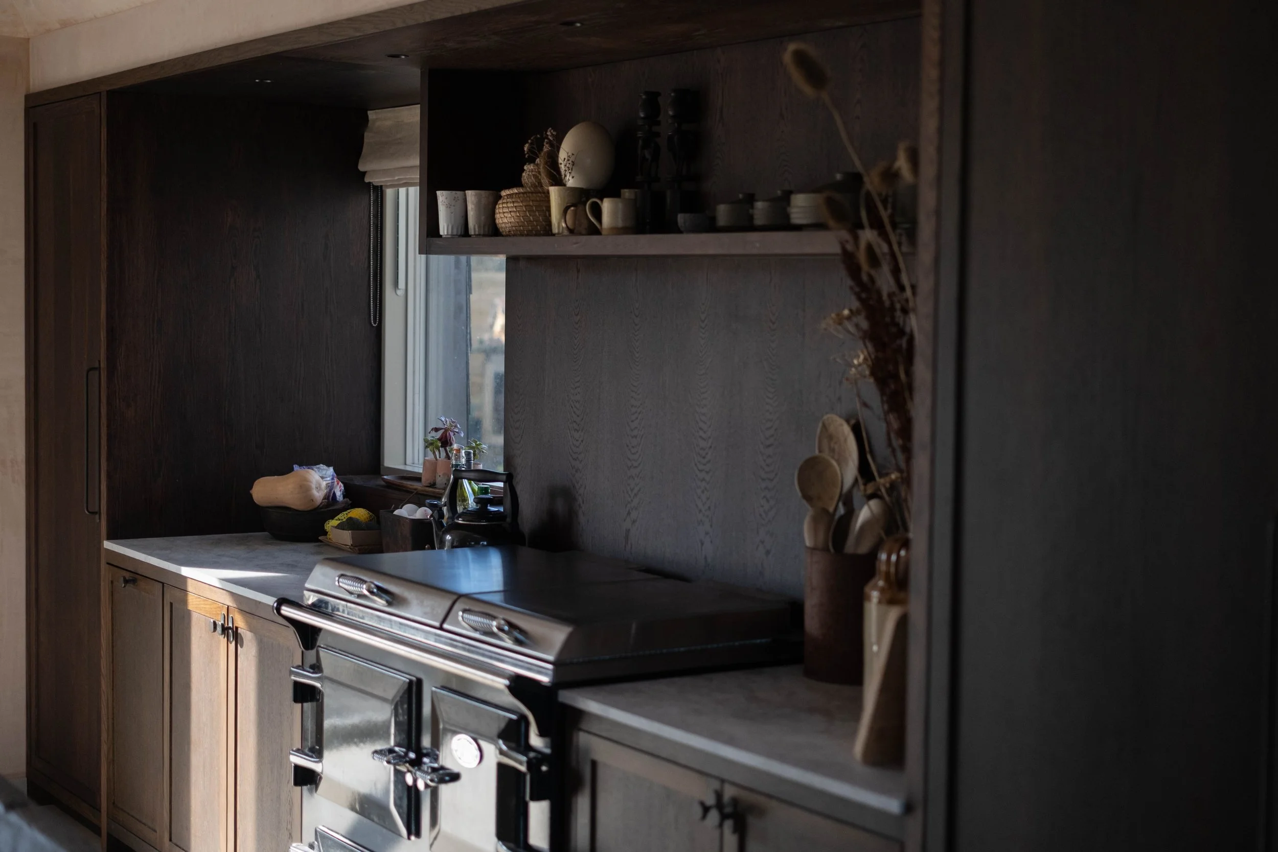 A kitchen with dark wood cabinets, a grey countertop, and a stainless steel oven. There are kitchen items, including a pumpkin, a plant, and various containers on the counter and on open shelves, with a window in the background letting in natural lig