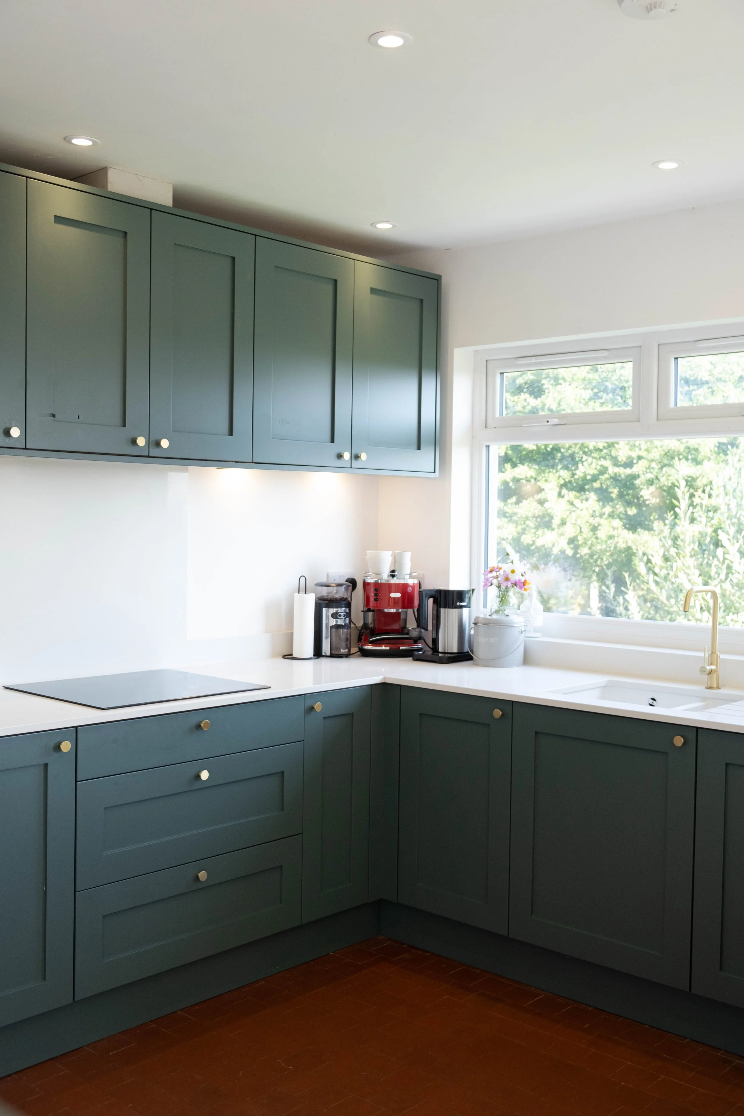 A kitchen with blue cabinets, white countertop, a window with greenery outside, and various small appliances on the counter including a coffee maker, kettle, and coffee machine.
