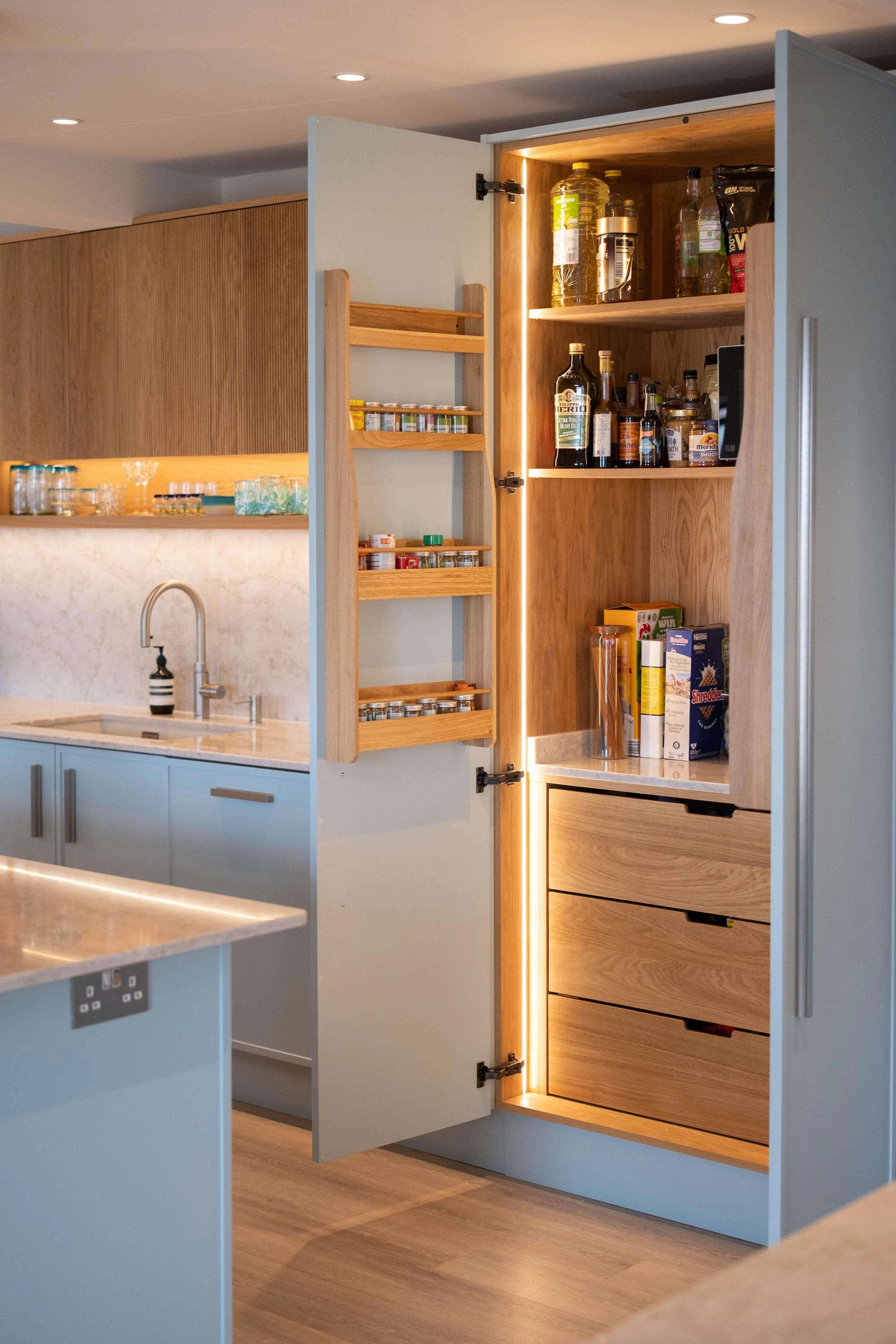 Open kitchen cabinet with wooden interior, shelves stocked with bottles of oil, sauces, and jars, illuminated with interior lighting, in a modern kitchen with light-colored cabinets and marble countertops.
