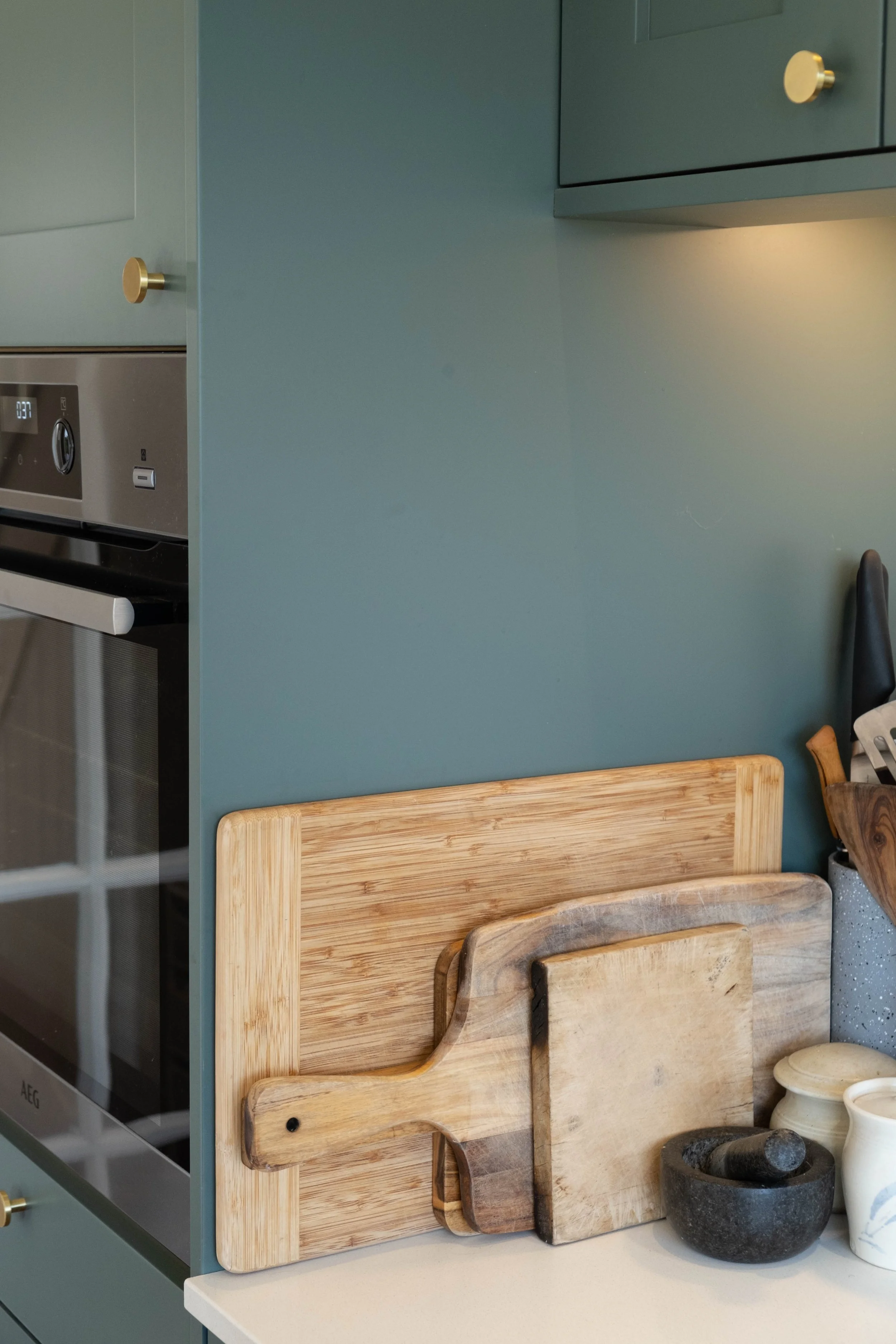 A kitchen countertop with a stack of wooden cutting boards with different shapes and sizes leaning against a teal wall. There is a black mortar and pestle, a white ceramic container, and a speckled black and white container on the counter.