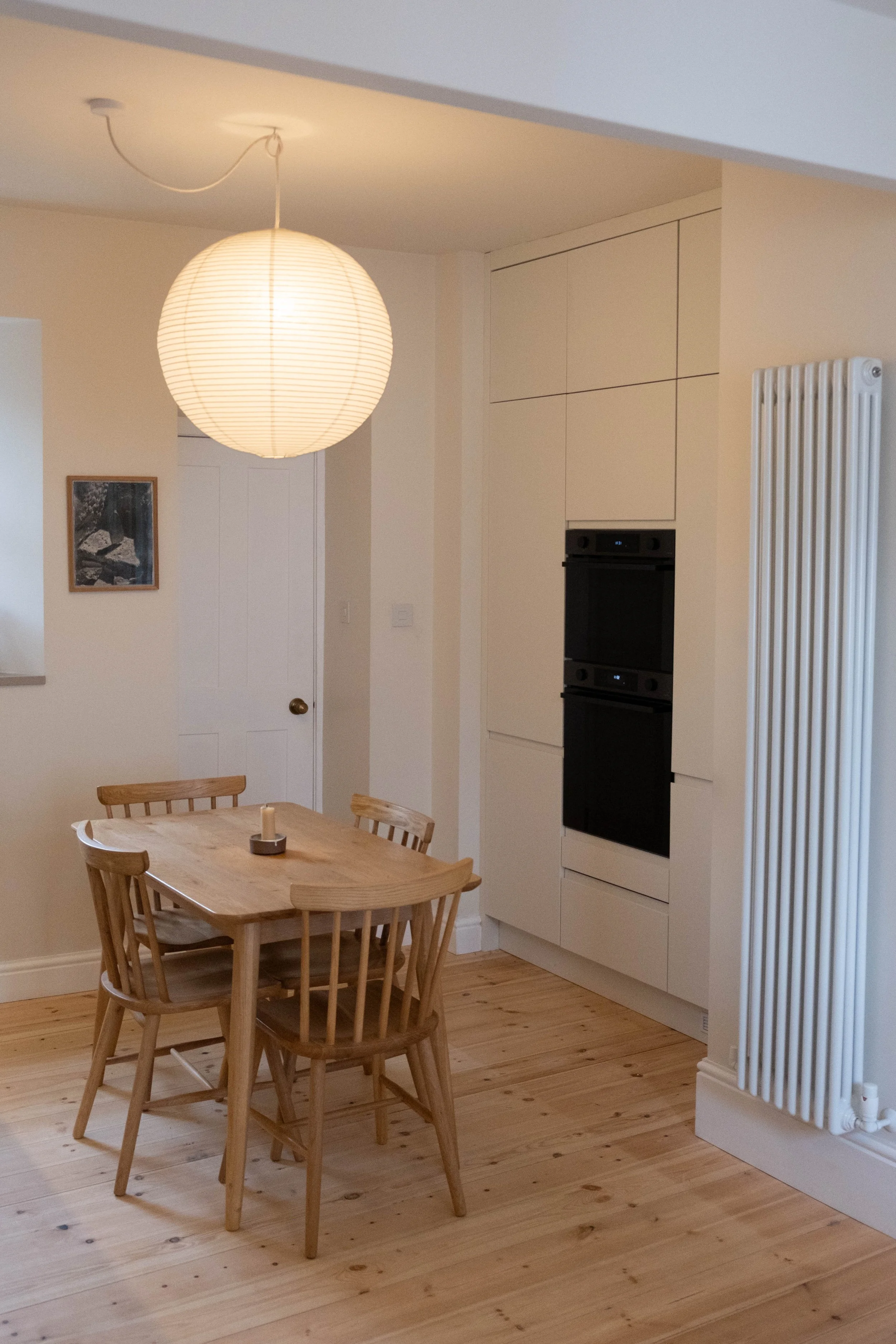 A dining area with a round wooden table surrounded by four wooden chairs, a white paper lantern pendant light hanging from the ceiling, a white wall with a small framed photo, a built-in oven in a cabinet, and a large white radiator beside the oven, 