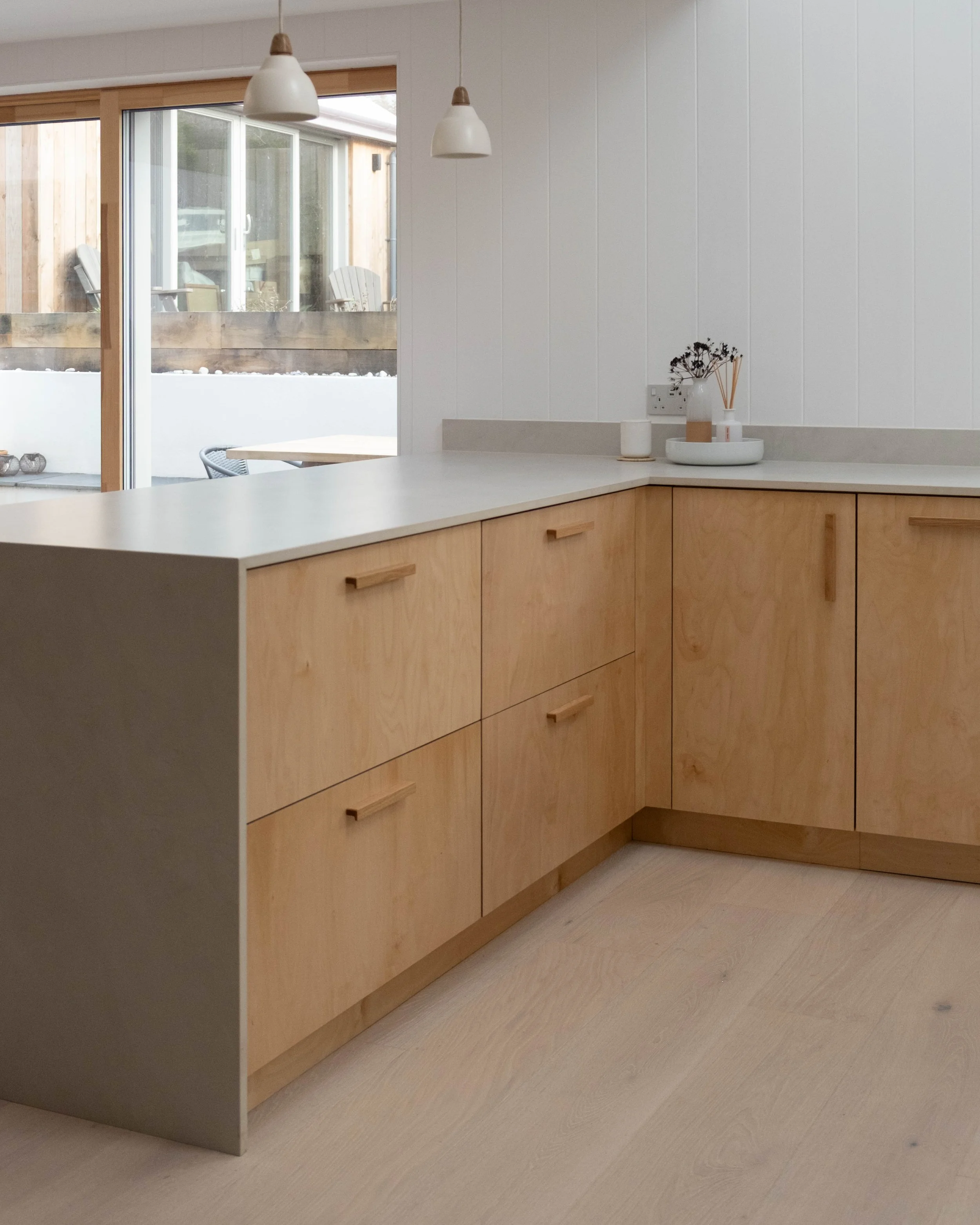 Interior view of a modern kitchen with beige wooden cabinets and a white countertop, decorated with a vase of dried flowers and reed diffuser, near a large sliding glass door leading to an outdoor patio area.