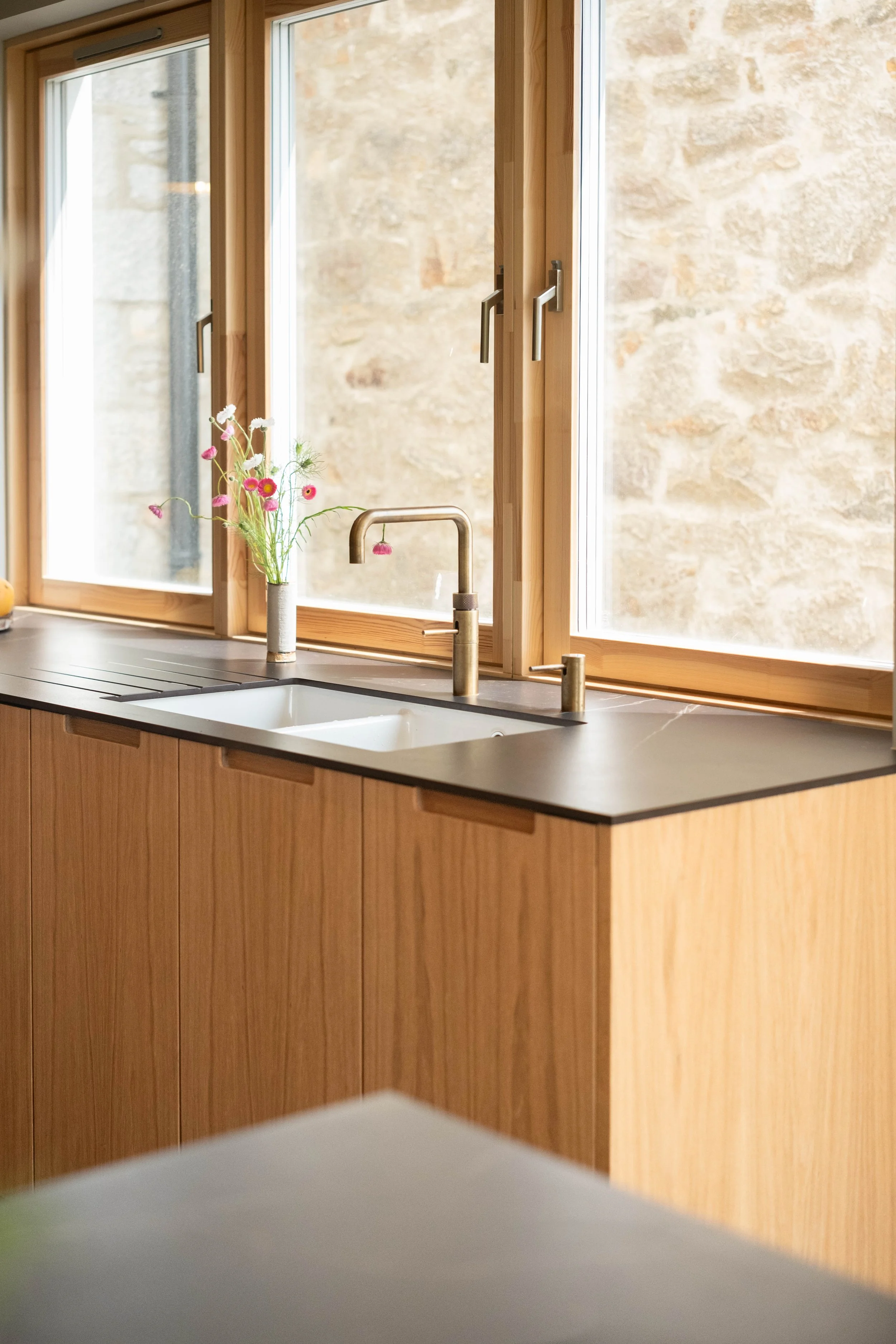 Kitchen window with wooden framing, a gold faucet, a vase of pink and white flowers, and a view of a stone wall outside.