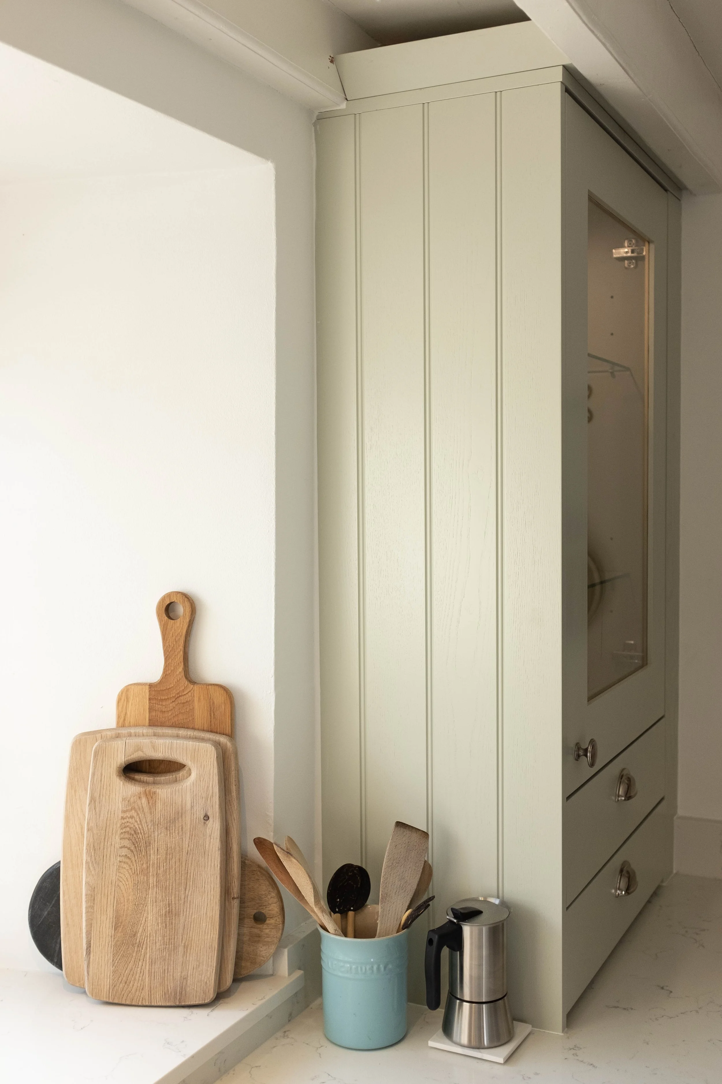 Kitchen countertop with wooden cutting boards, utensils in a blue container, and a silver coffee percolator. Light green cabinets are in the background.