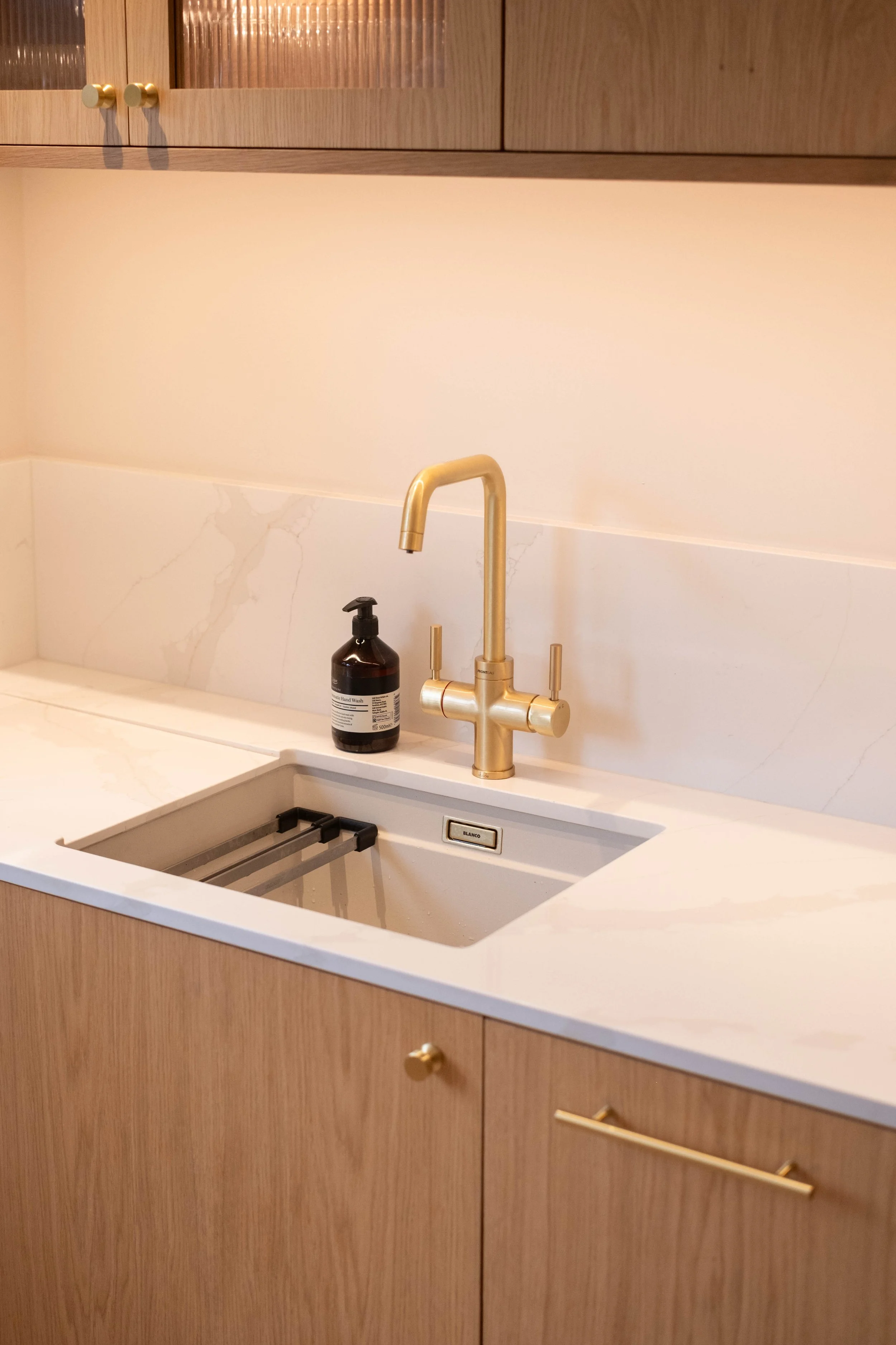 A kitchen sink with a gold-colored faucet, a black soap dispenser, and wooden cabinetry with gold knobs.