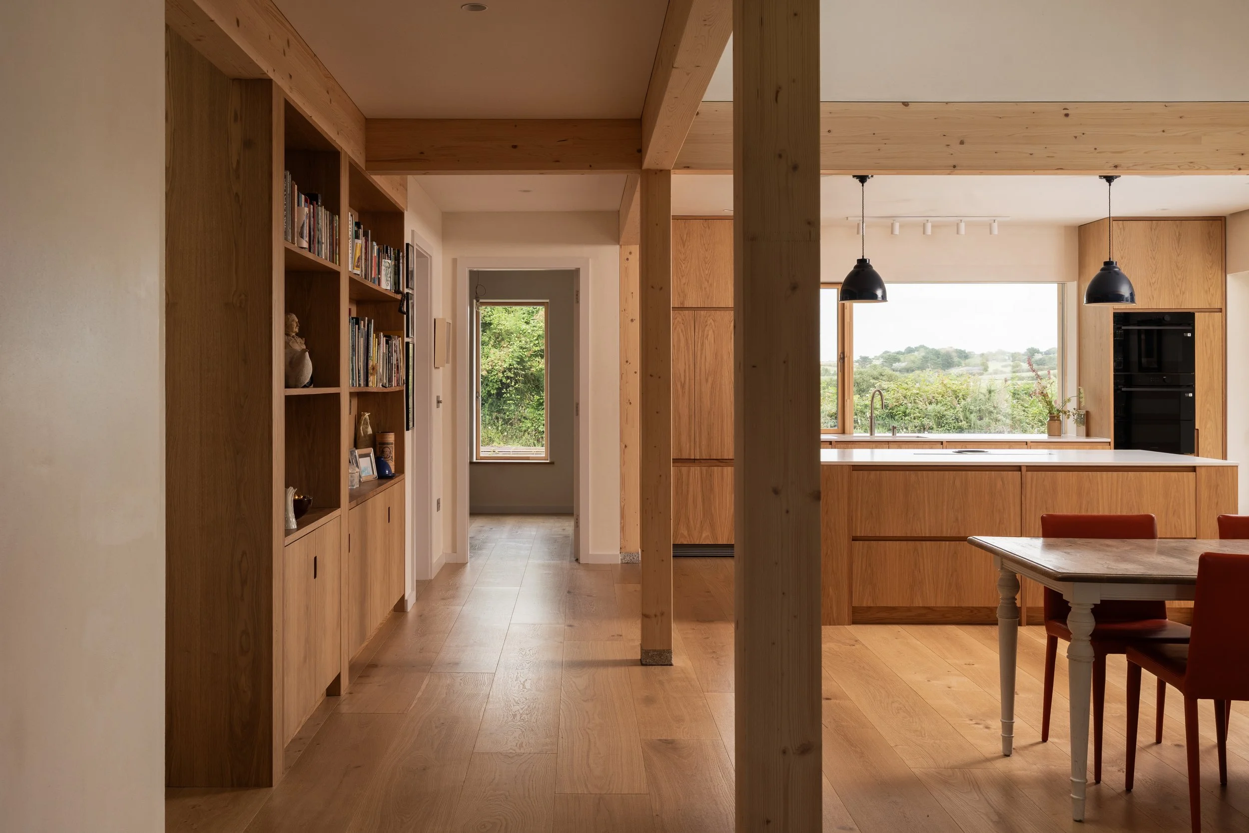 Interior of a modern kitchen with wooden cabinets, large window overlooking greenery, and a dining table with red chairs.