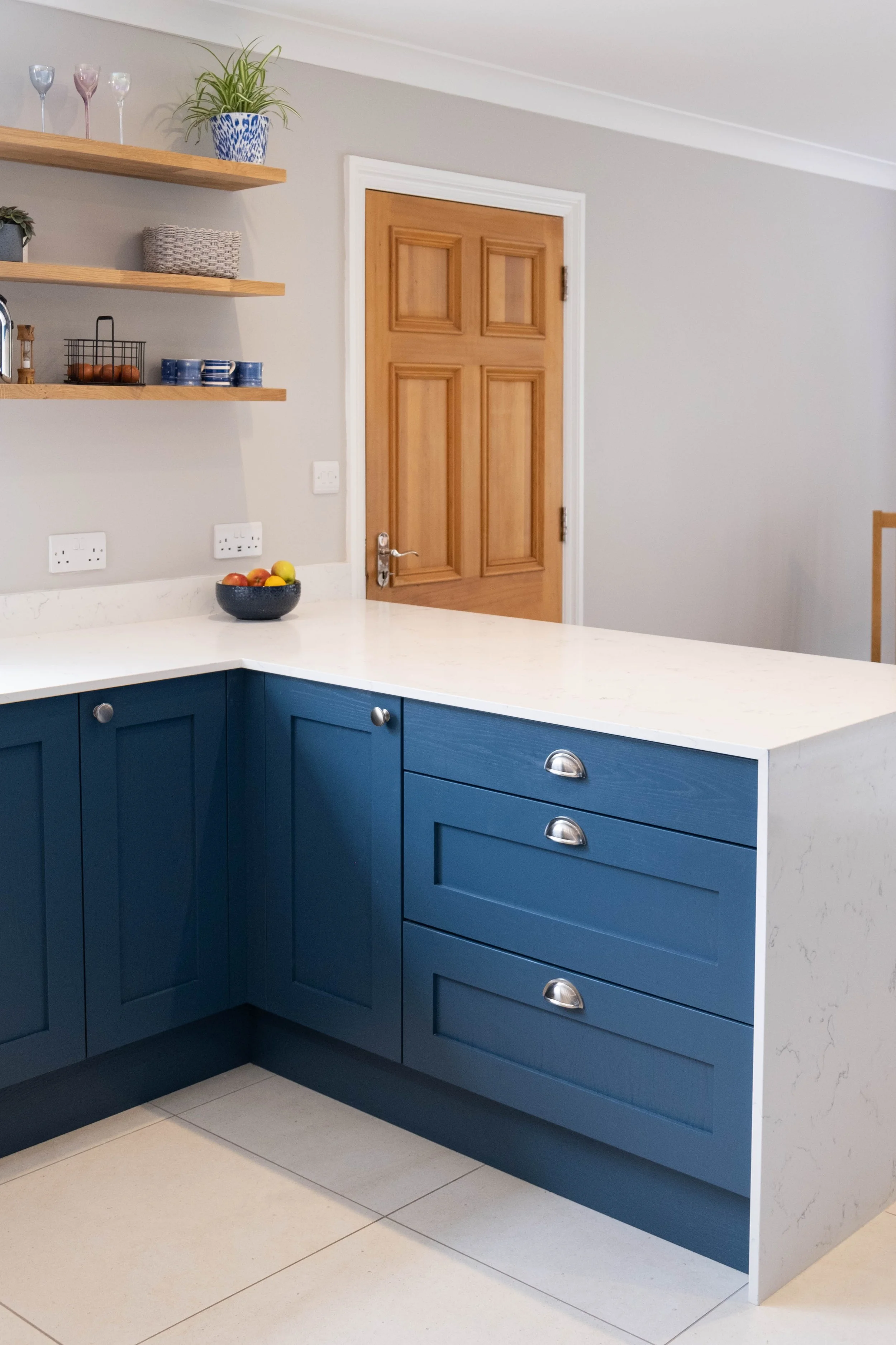 Kitchen with blue cabinets, white countertop, wooden door, white tiled floor, wall shelves with plants and glassware, black fruit bowl with apples, electrical outlets on the wall.