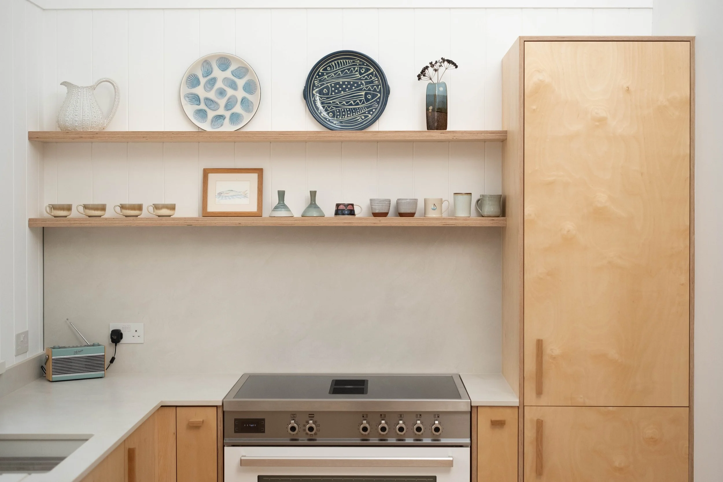 Kitchen with wooden cabinetry, white walls, open shelving displaying decorative plates, vases, and cups, and a stovetop with control knobs. There is a small heater and electrical outlet on the lower wall.