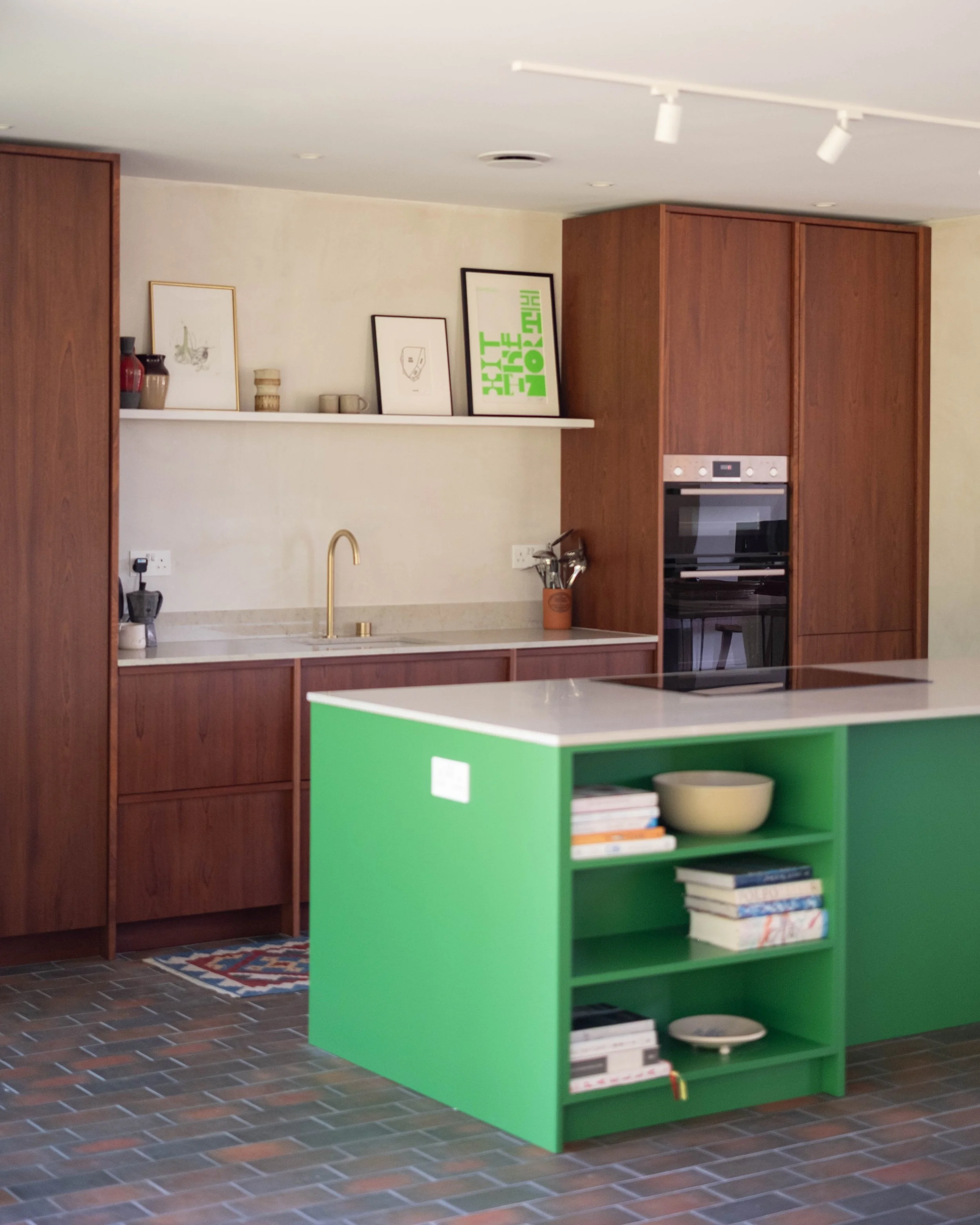 A kitchen with a wooden cabinet, a white countertop with a gold faucet, a small green bookshelf holding books and dishes, framed artwork on a white shelf, and a tiled floor.