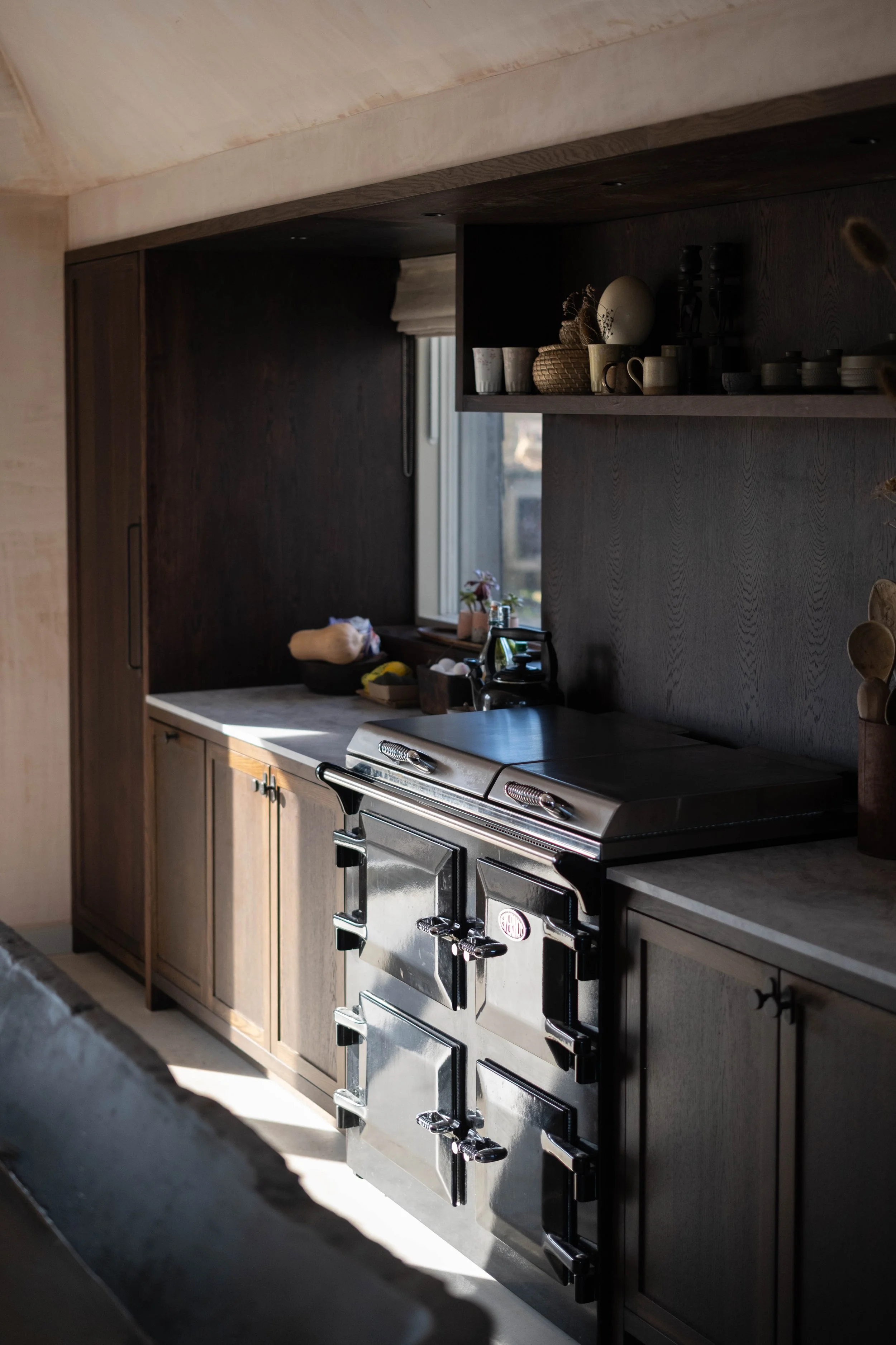A kitchen with dark wood cabinets, a stainless steel oven with black accents, and a countertop with various items including a basket, mugs, and a kettle. Sunlight illuminates part of the room through a window.