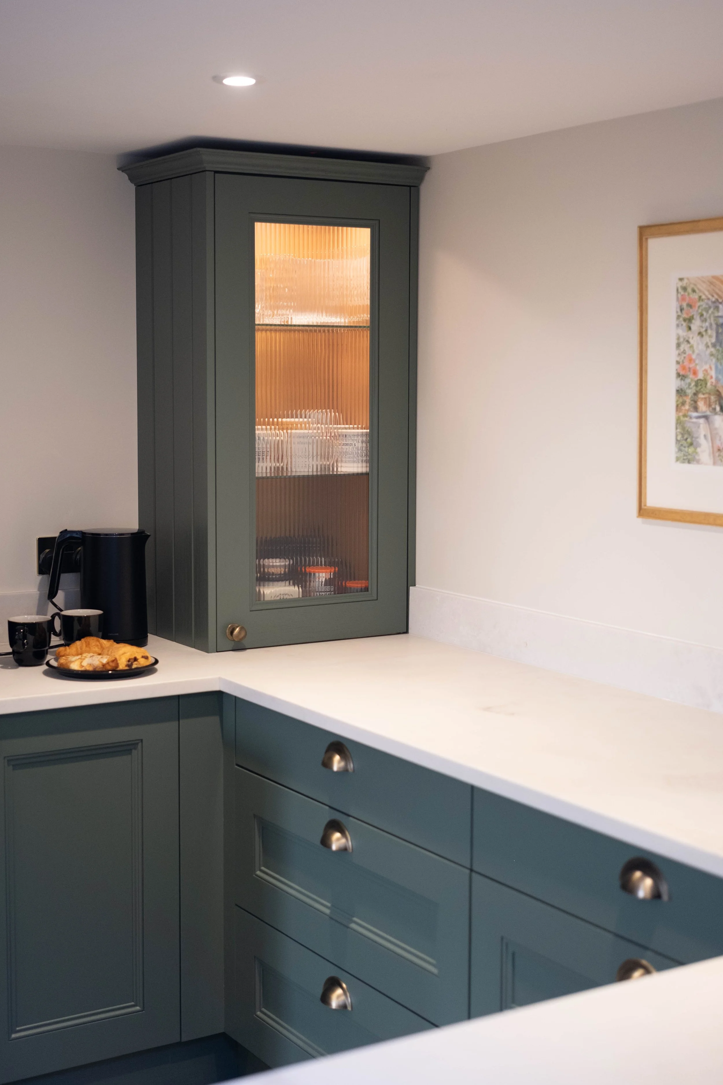 A kitchen with blue cabinets, a white marble countertop, a framed picture on the wall, and a glass-fronted cabinet with baked goods and cups on the counter.
