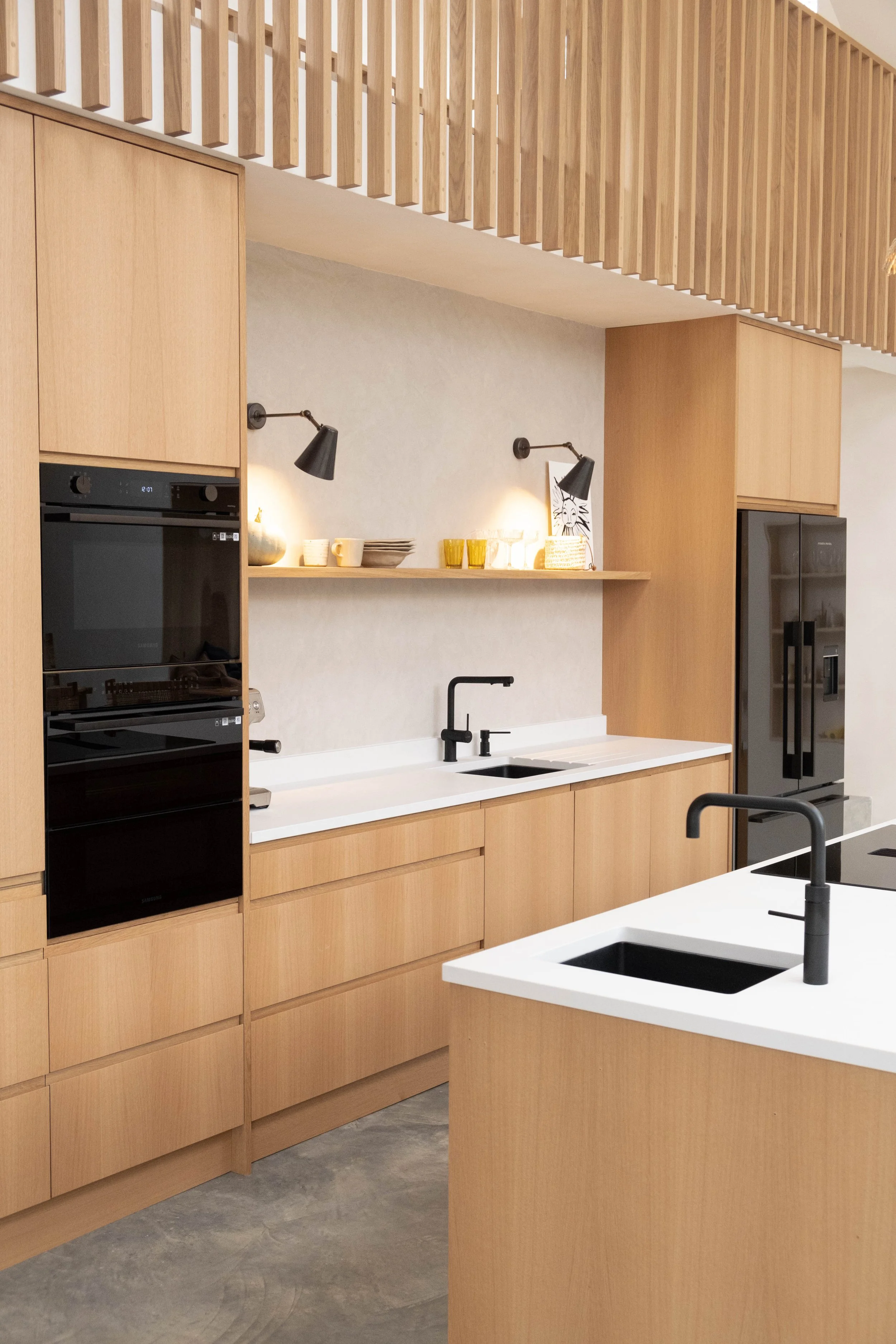 Modern kitchen with light wood cabinets, white countertops, black sinks, and black wall-mounted lamps. A shelf with cups and plates, and a partial view of a refrigerator and oven.