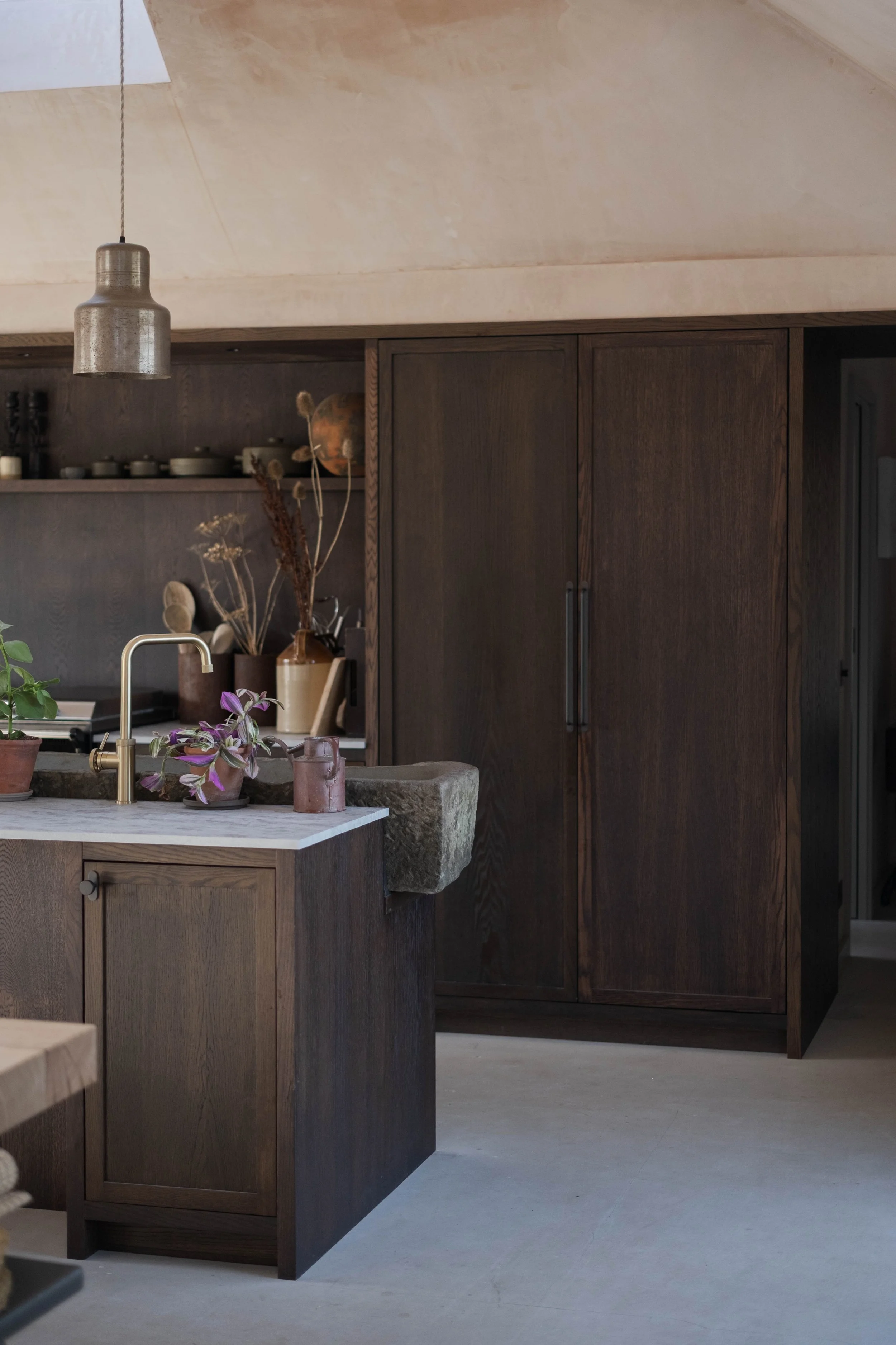 A modern kitchen featuring a wooden island with a white marble countertop, decorated with potted plants and dried flowers, with dark wooden cabinets and open shelving in the background.