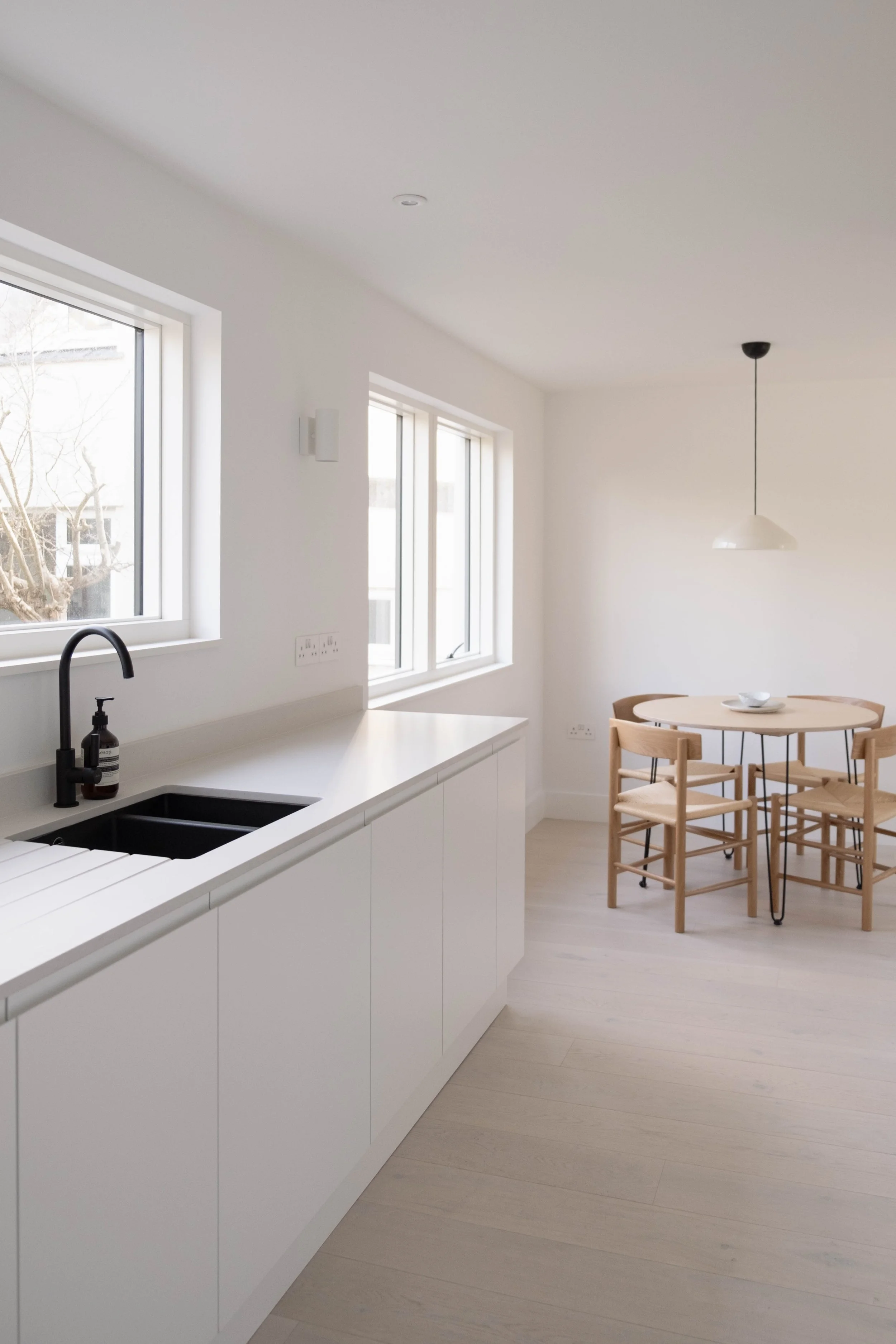 A bright, minimalist kitchen with white cabinets, a white countertop, a black faucet, and a window showing trees outside. Adjacent to the kitchen is a dining area with a round wooden table, four chairs, and a white pendant light hanging from the ceil