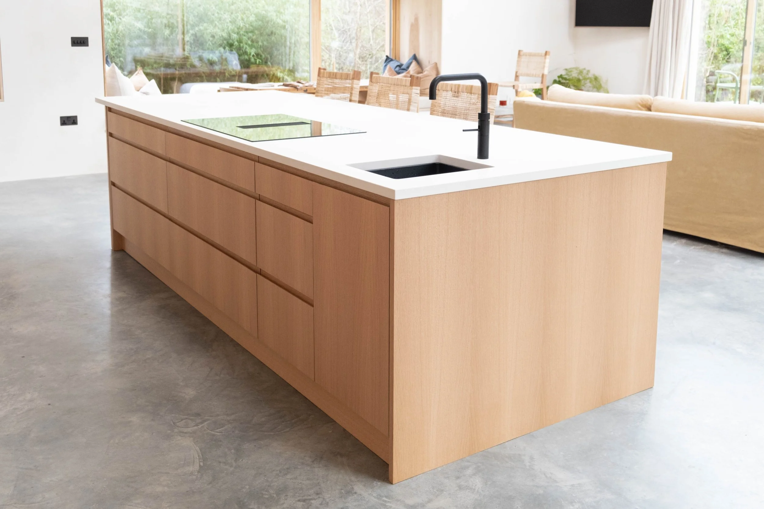Modern kitchen island with a black faucet, white countertop, and light wood cabinetry, in an open-concept living space with large windows and outdoor greenery.