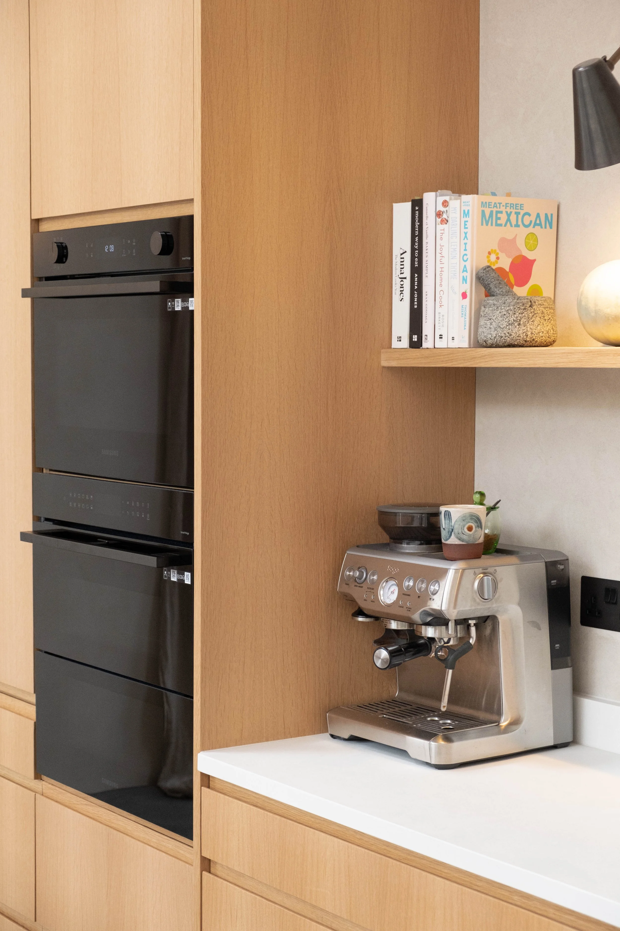 A modern kitchen with a built-in oven, a coffee machine, a small shelf with cookbooks, and a lamp with a globe-shaped shade.