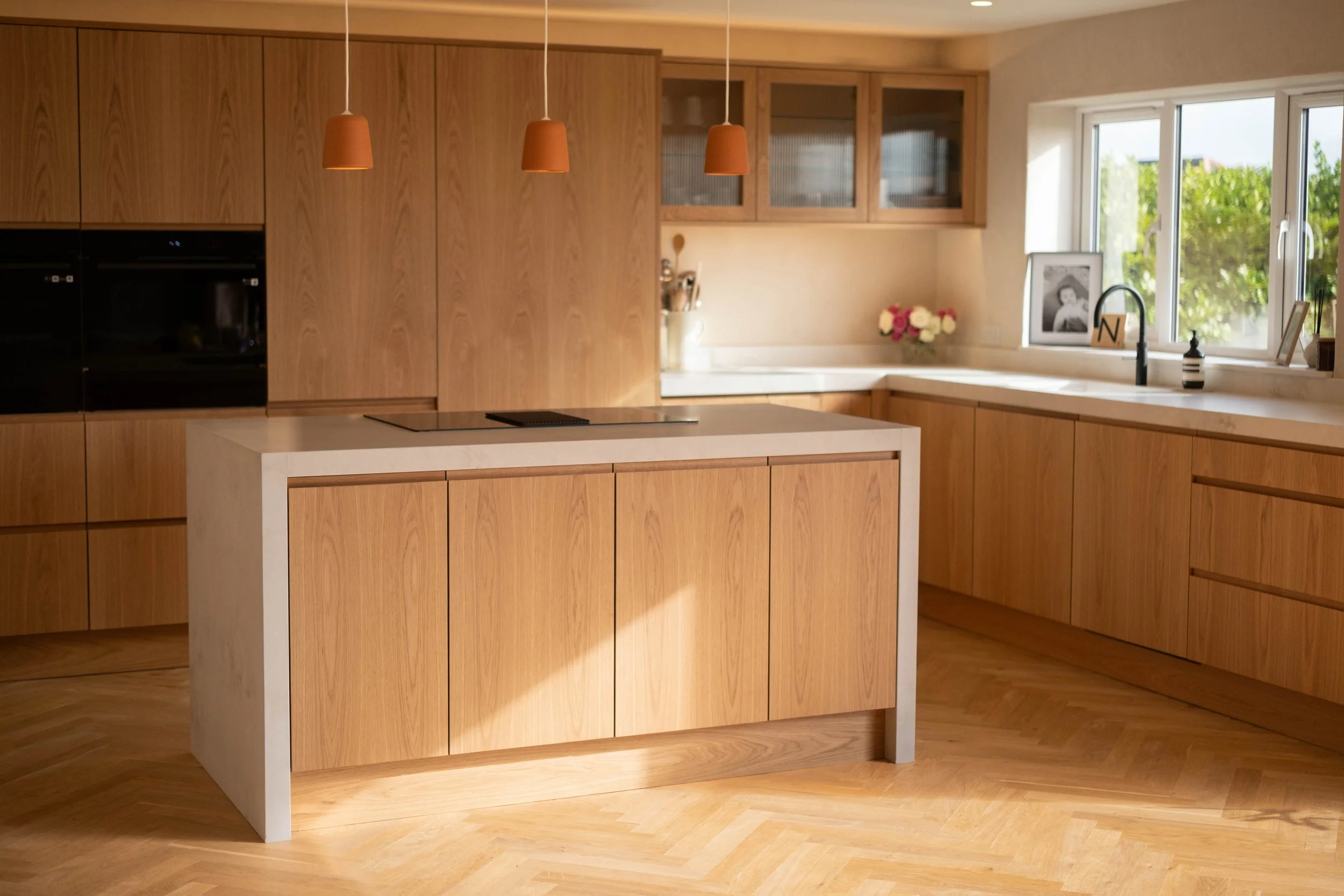 Modern kitchen with wooden cabinets, a white kitchen island, a large window with a view of greenery, and pendant lights hanging from the ceiling.