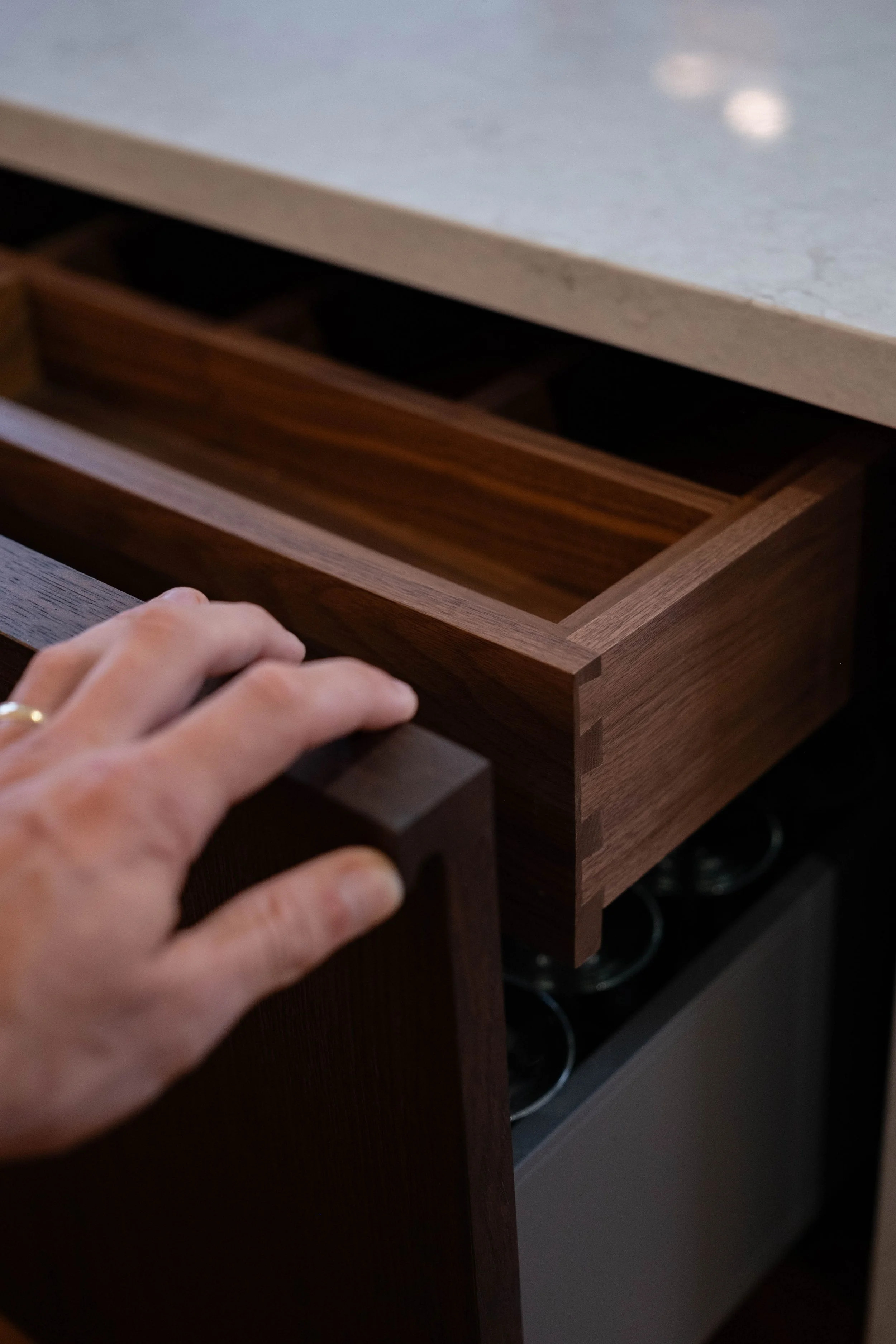 A hand opening a wooden kitchen drawer with slotted compartments, beneath a light-colored stone countertop.