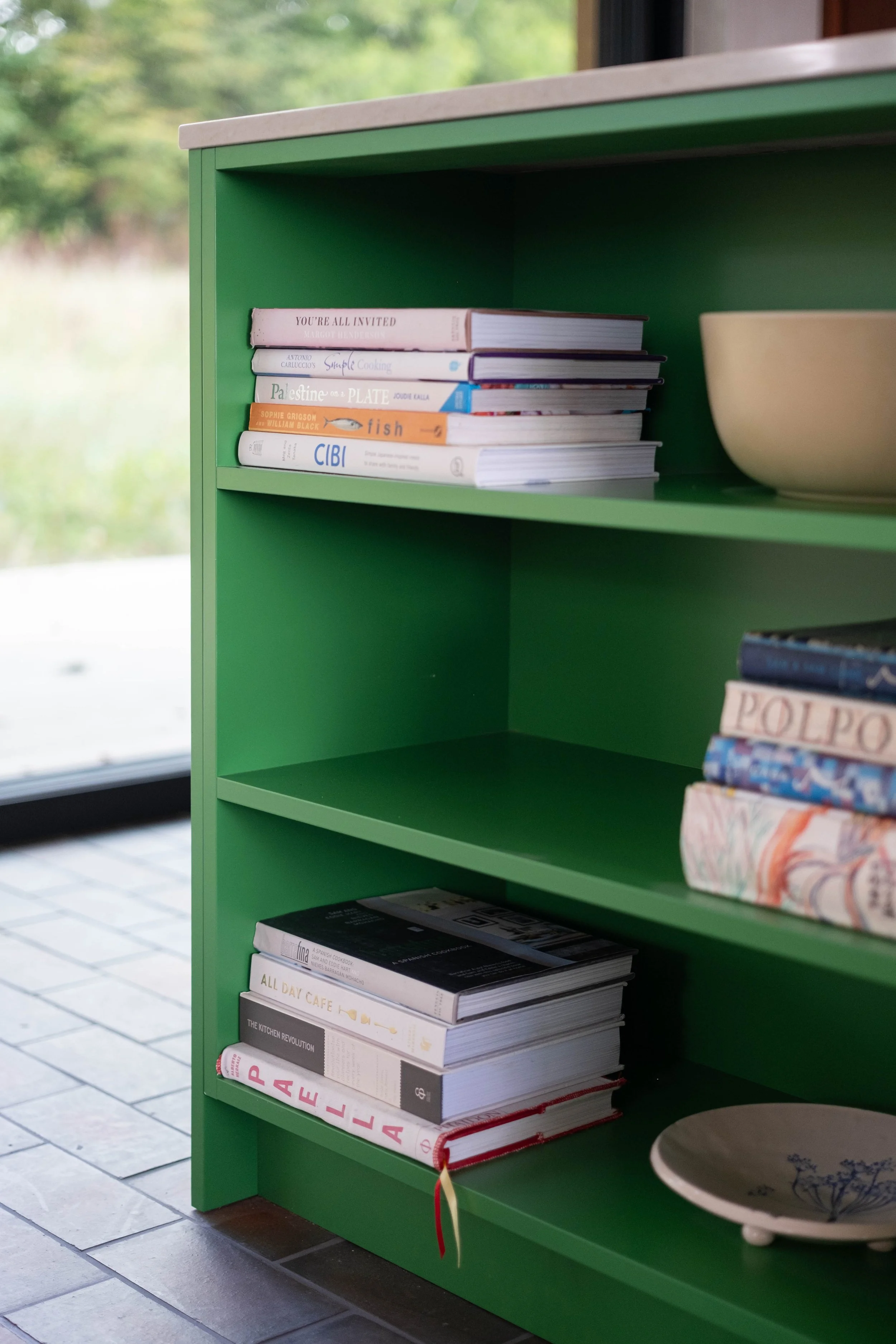 A green bookshelf next to a window, holding various stacked books and a ceramic bowl with a floral design.
