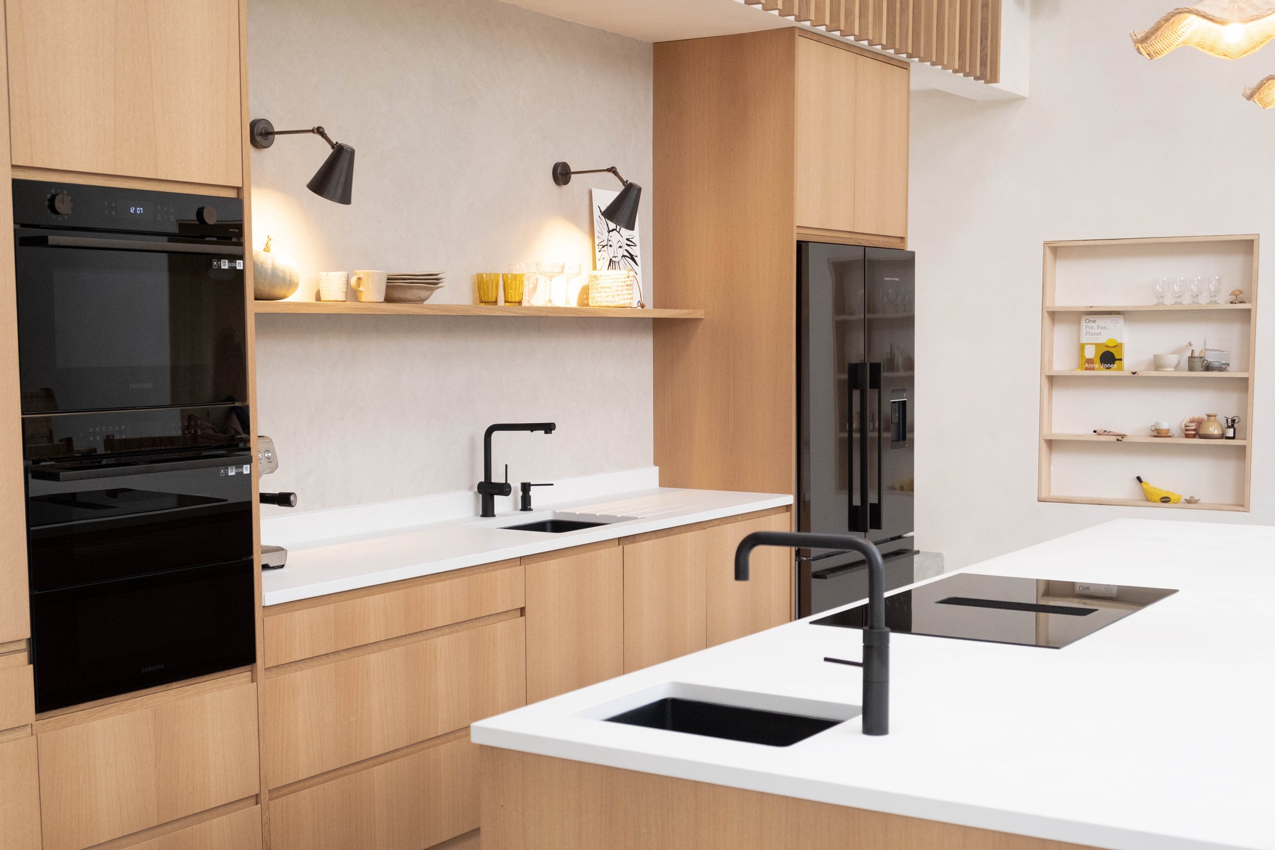 Modern kitchen with light wood cabinetry, white countertops, black sinks, black wall-mounted lamps, and a black refrigerator.