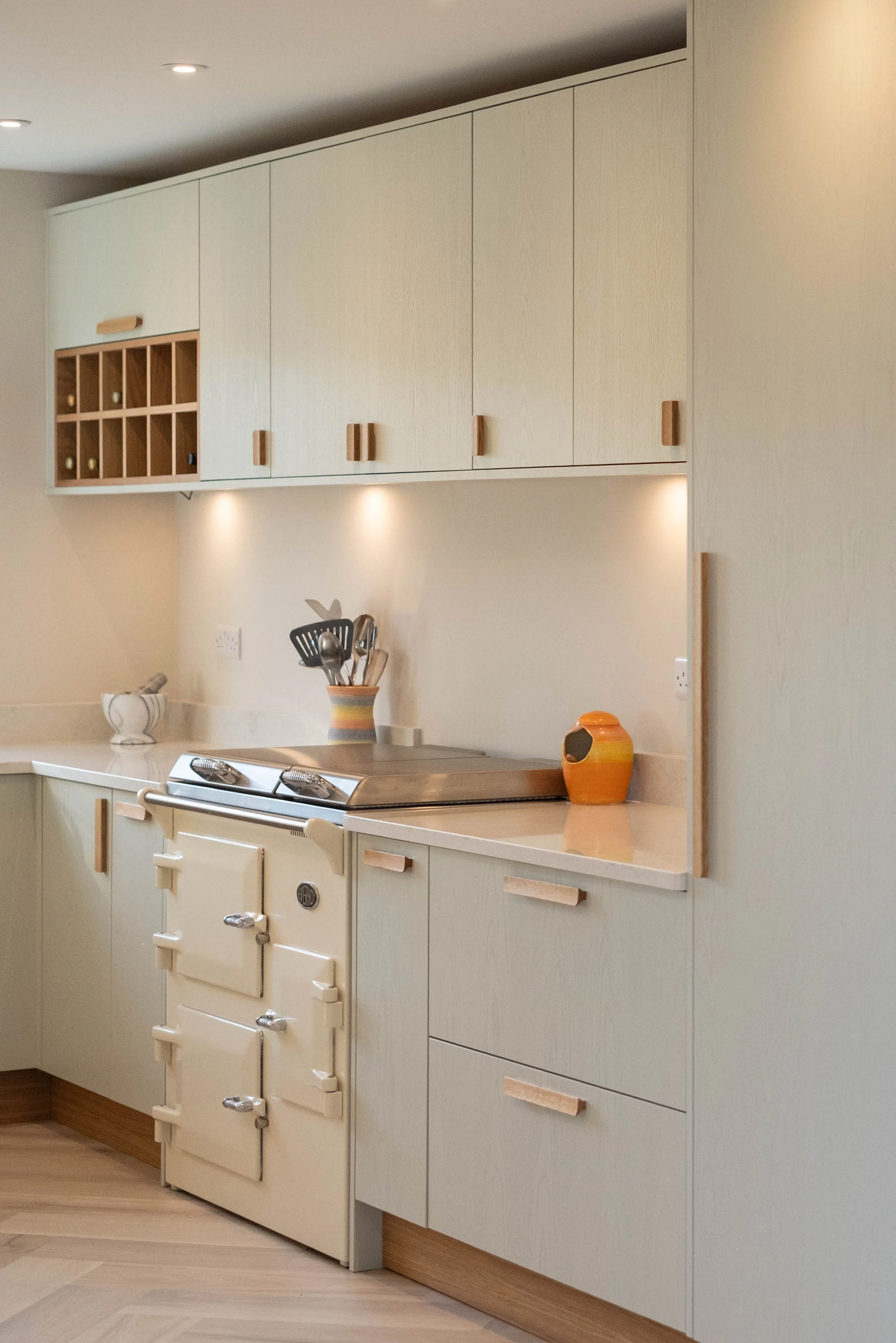 A kitchen with beige cabinets and drawers, a vintage stove, a countertop with a cup holding kitchen utensils, and decorative vases, with recessed ceiling lights.