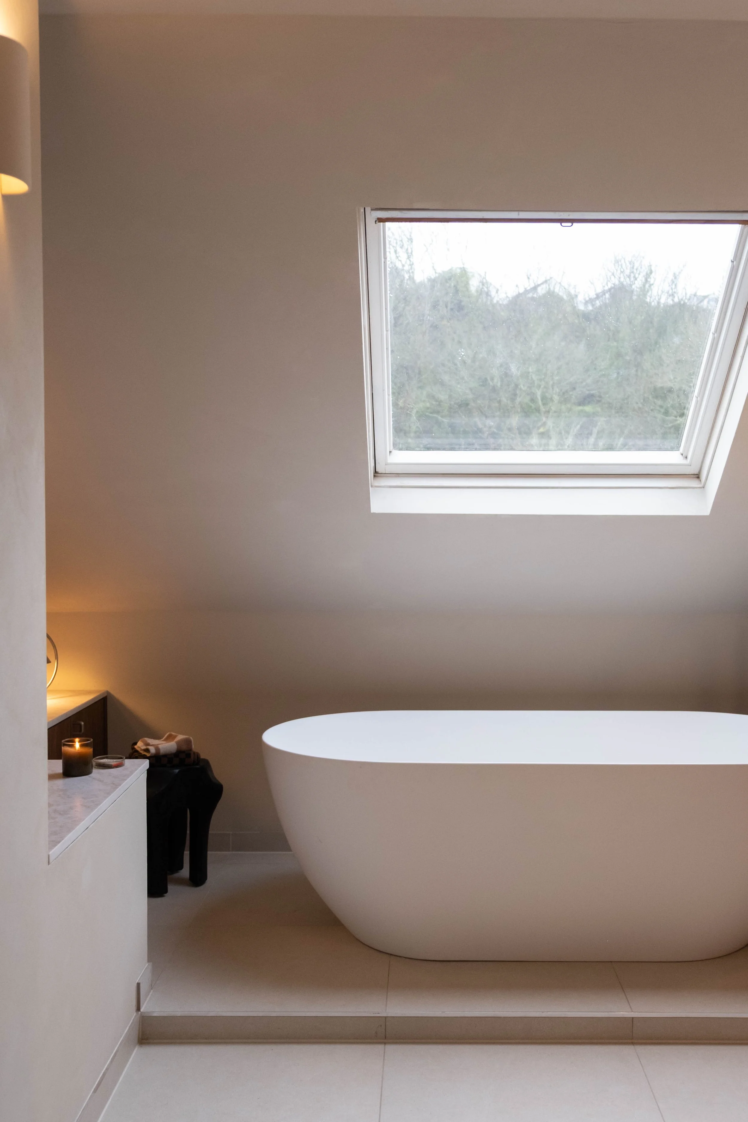 A bathroom with a view of a large skylight window, a modern freestanding bathtub, and a side table with candles and toiletries.